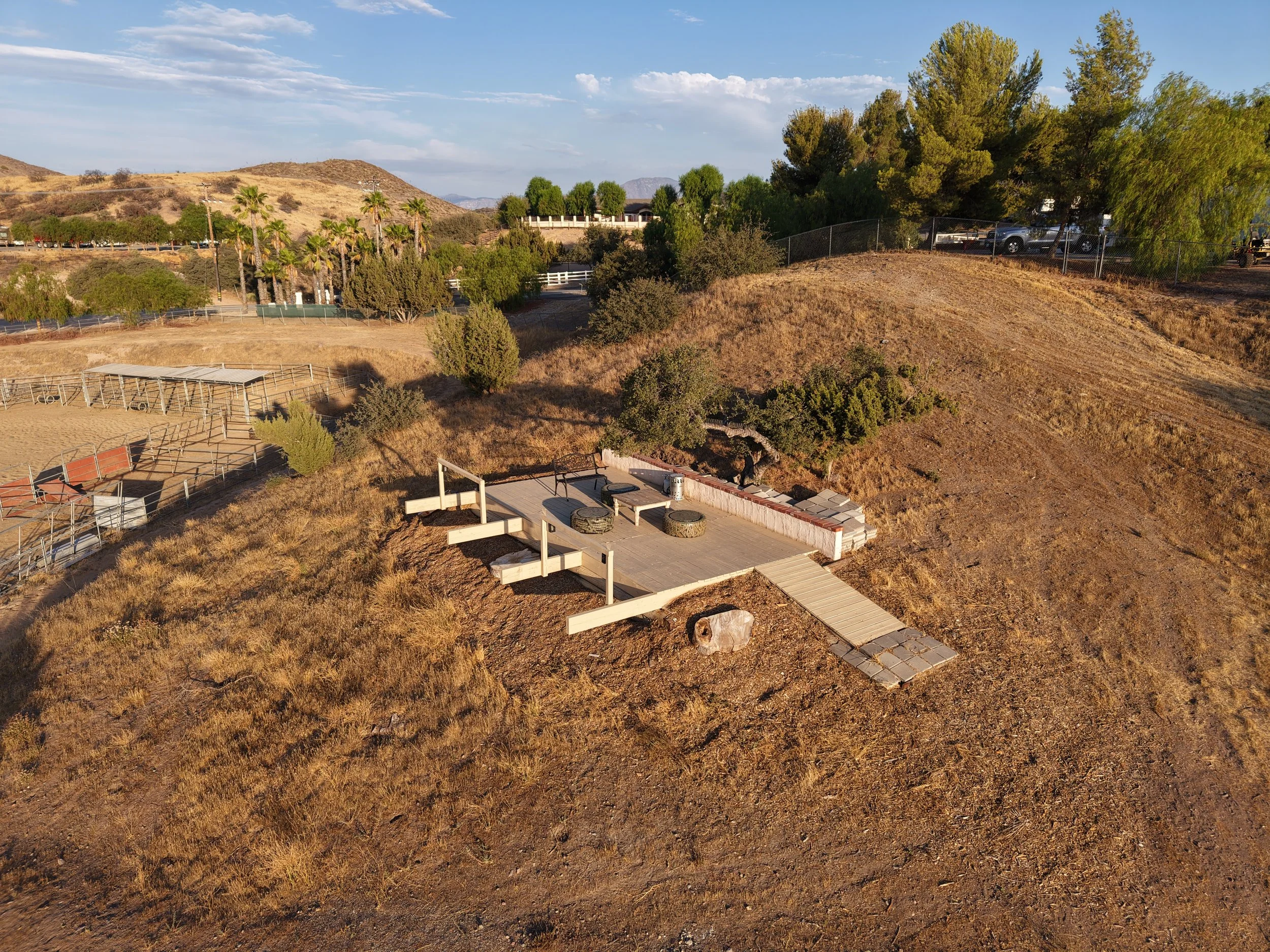 An aerial view of a dry, hilly landscape with a small, newly constructed wooden deck area featuring a few outdoor furniture pieces, including tires used as seats and a small table, with a brick walkway leading to it. Surrounding the area are sparse bushes and trees, with a section of fenced parking lot and more greenery in the background.
