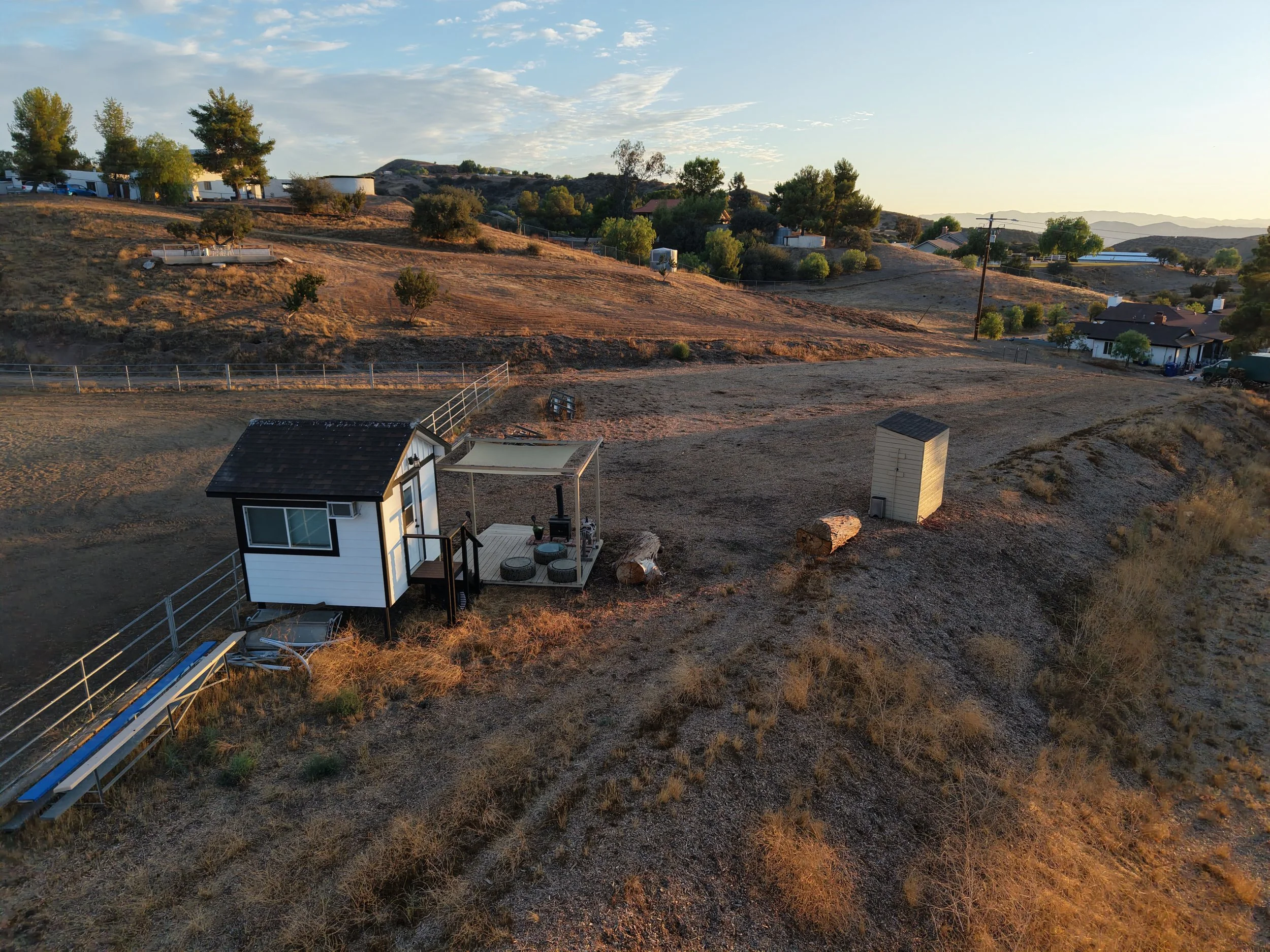 A residential backyard with a small white house with black trim, an outdoor seating area with cushions, a ramp, and a slide leading down a grassy hill. In the field, there are rocks, bushes, and a small shed. Other houses and trees are visible in the background, with hills and a sunset sky.