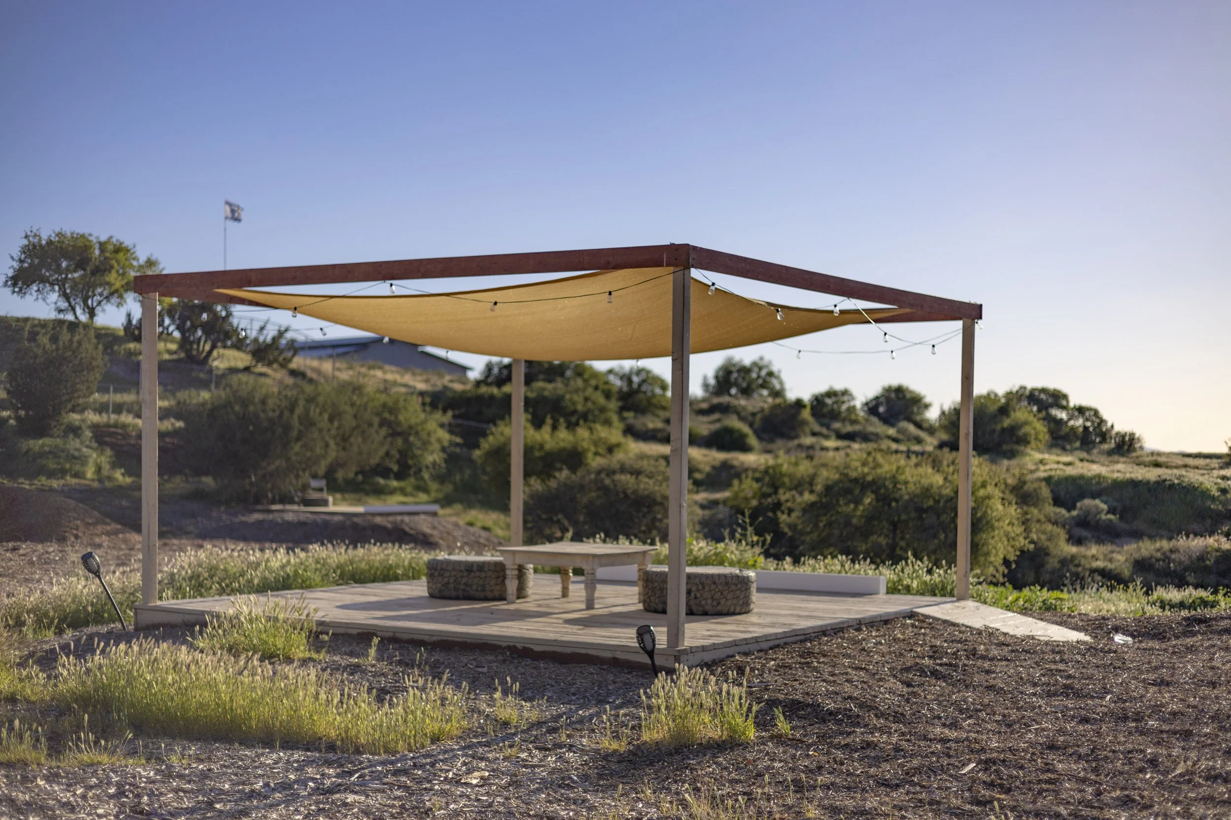 An outdoor wooden deck with a beige shade sail overhead, surrounded by natural landscape, with two round seats, a small table, and landscape lighting fixtures.