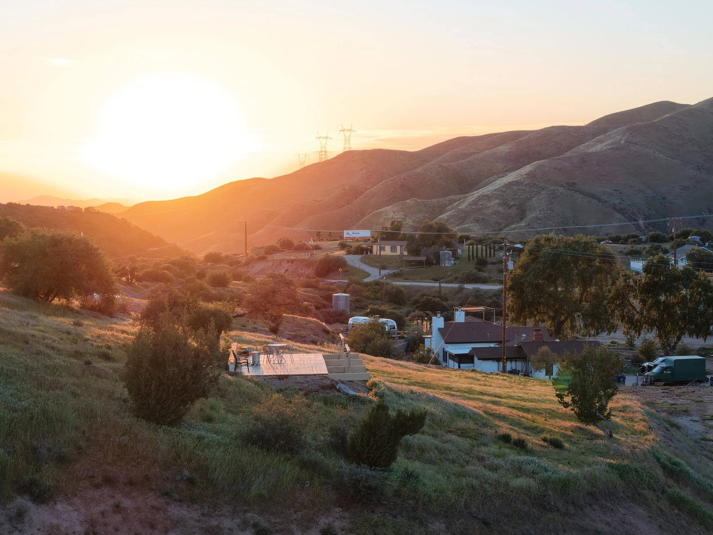 Sunset over a rural landscape with rolling hills, scattered trees, a few houses, and power lines