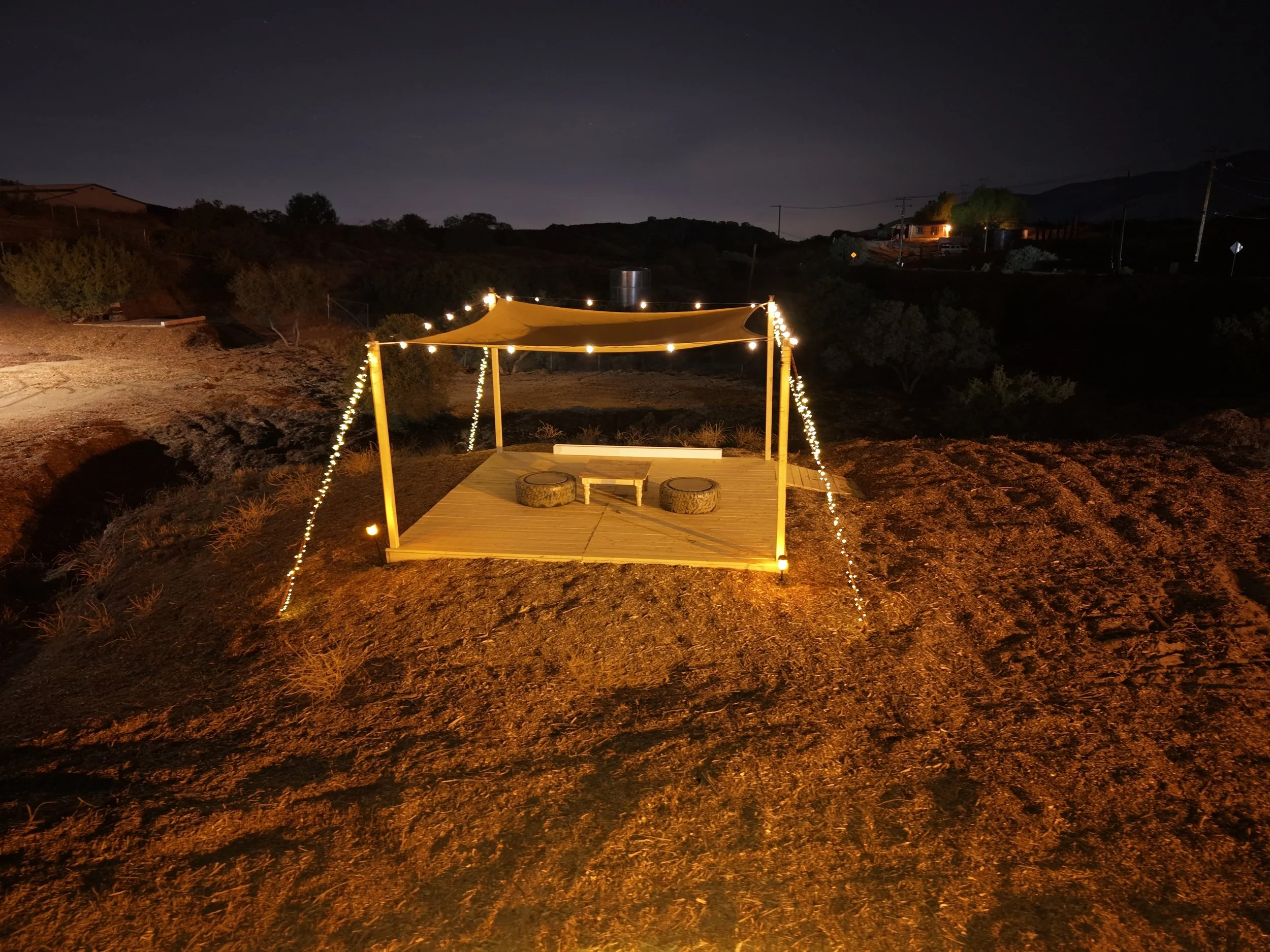 Nighttime outdoor scene with a small wooden platform under a canopy, decorated with string lights, and two logs serving as seats, placed on a dirt hill with some trees and distant houses in the background.