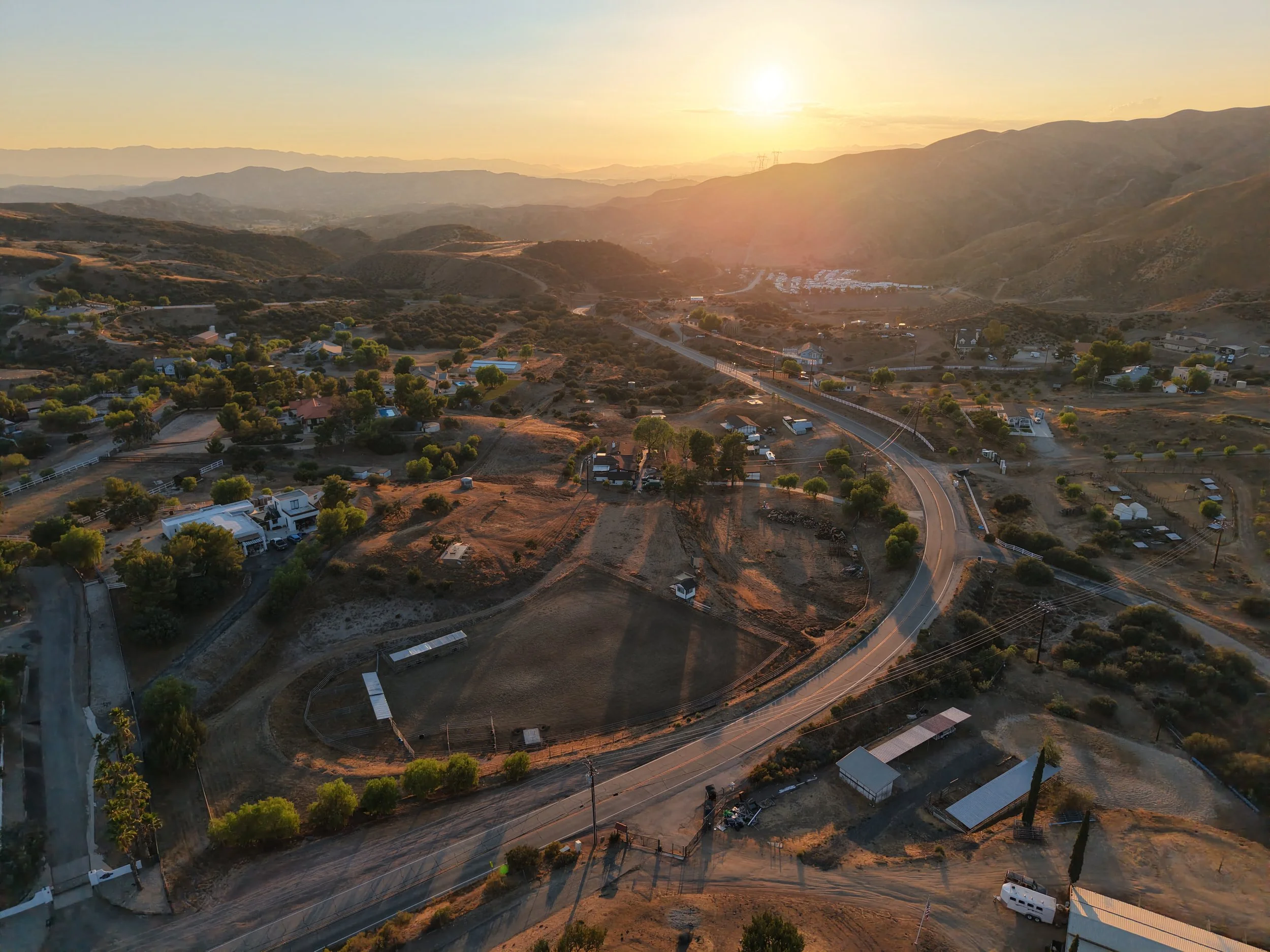 An aerial view of a rural landscape at sunset, showing winding roads, scattered houses, trees, and hills with mountains in the distance.