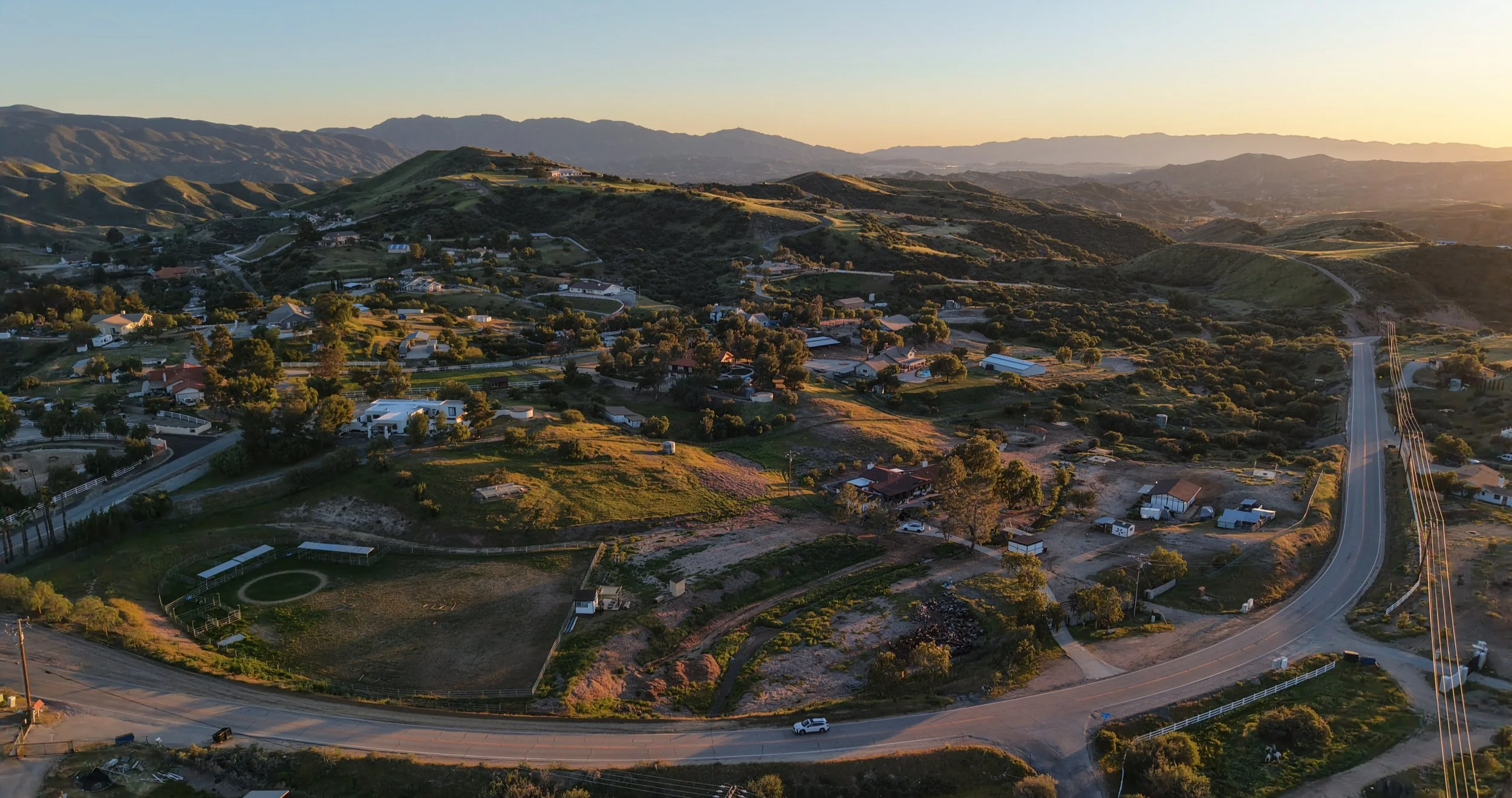 Aerial view of a rural hillside neighborhood with houses, trees, and winding roads during sunset.