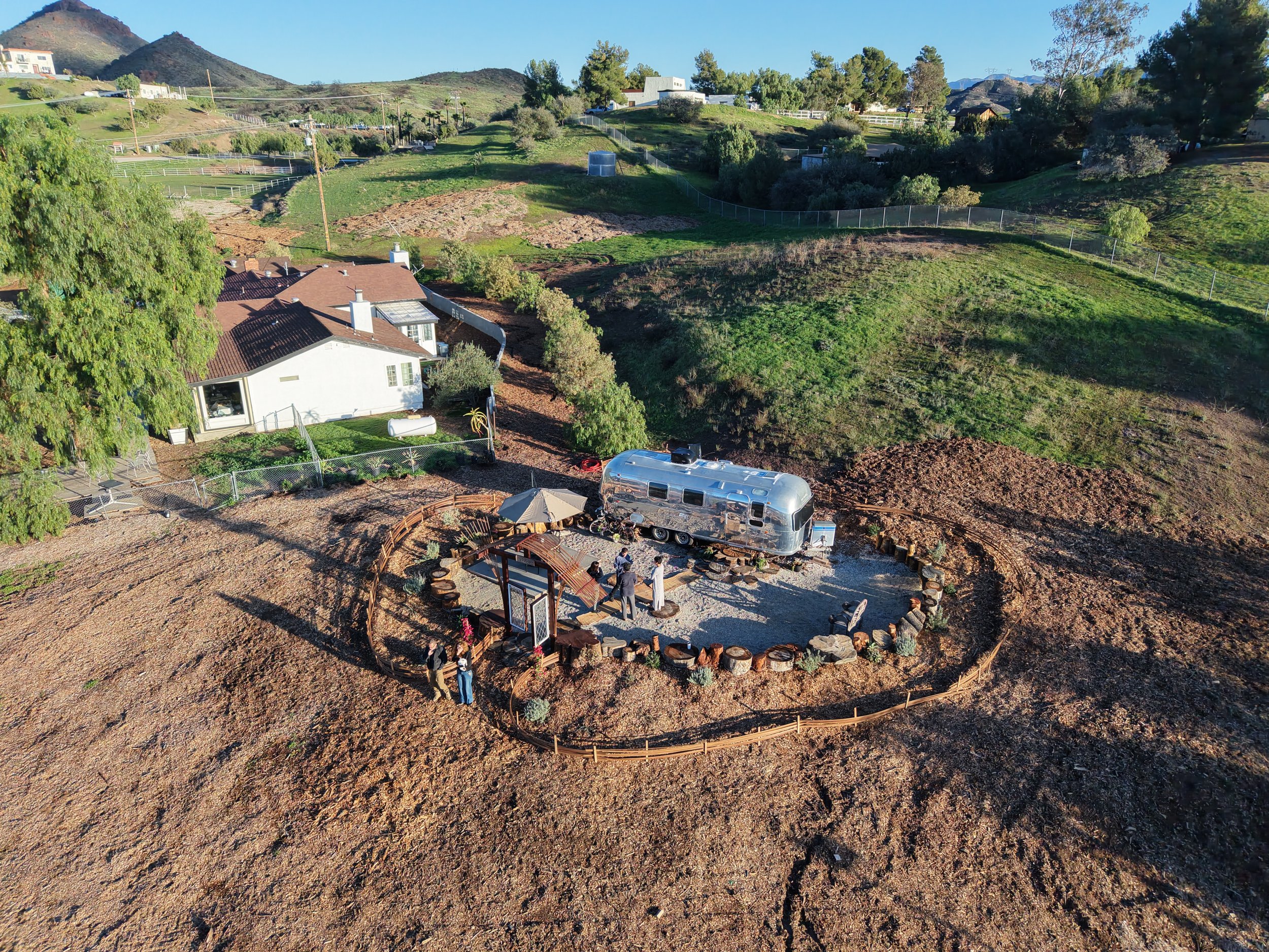 An aerial view of a backyard with a shiny metallic Airstream trailer, outdoor seating area with a table, chairs, and a large umbrella, surrounded by a circular border of logs and rocks, located in a semi-arid landscape with neighboring houses and green hills in the background.
