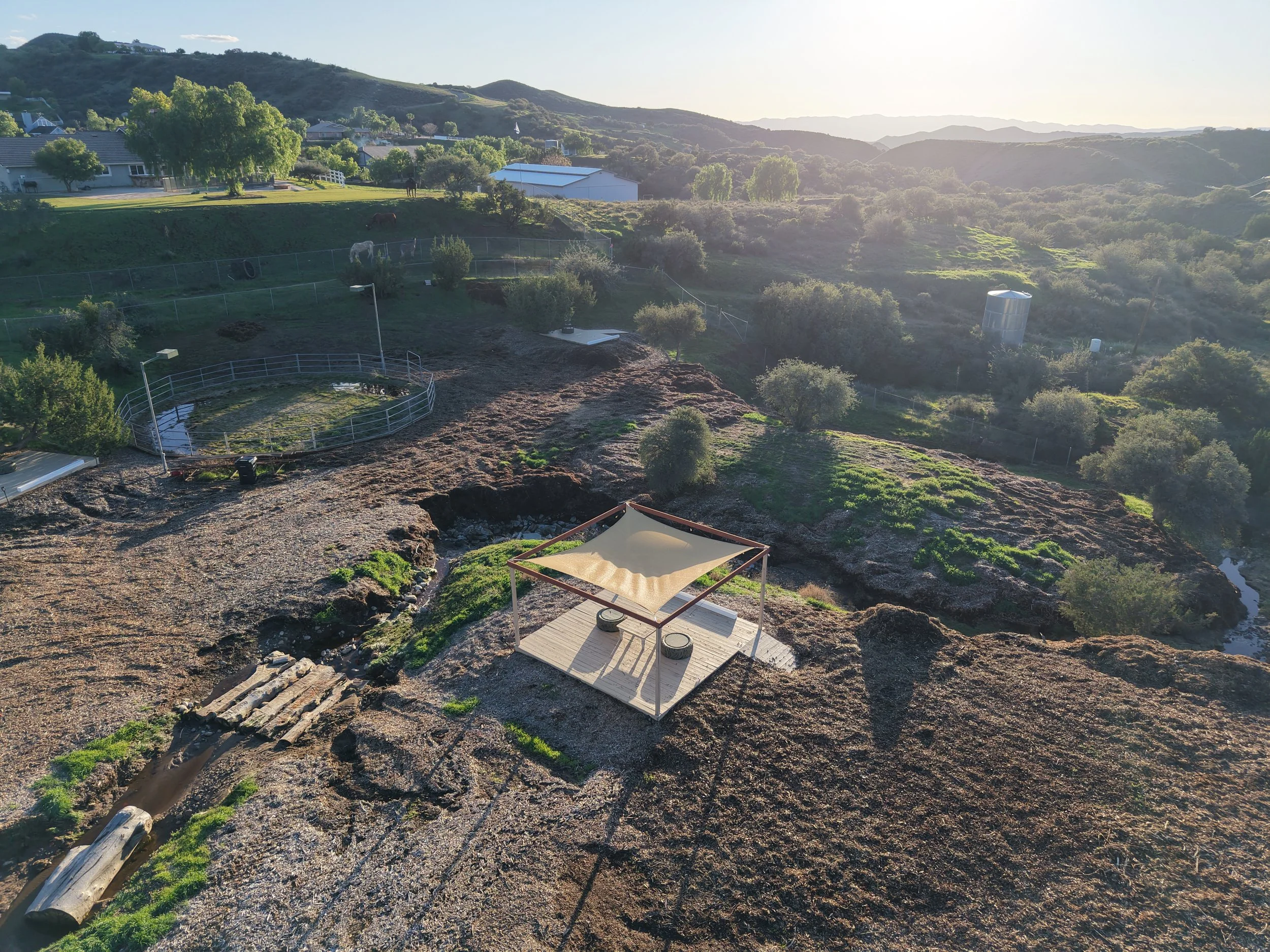 A serene rural landscape with rolling hills, trees, and an outdoor shaded seating area with a canopy, with a small pond and rocky terrain.