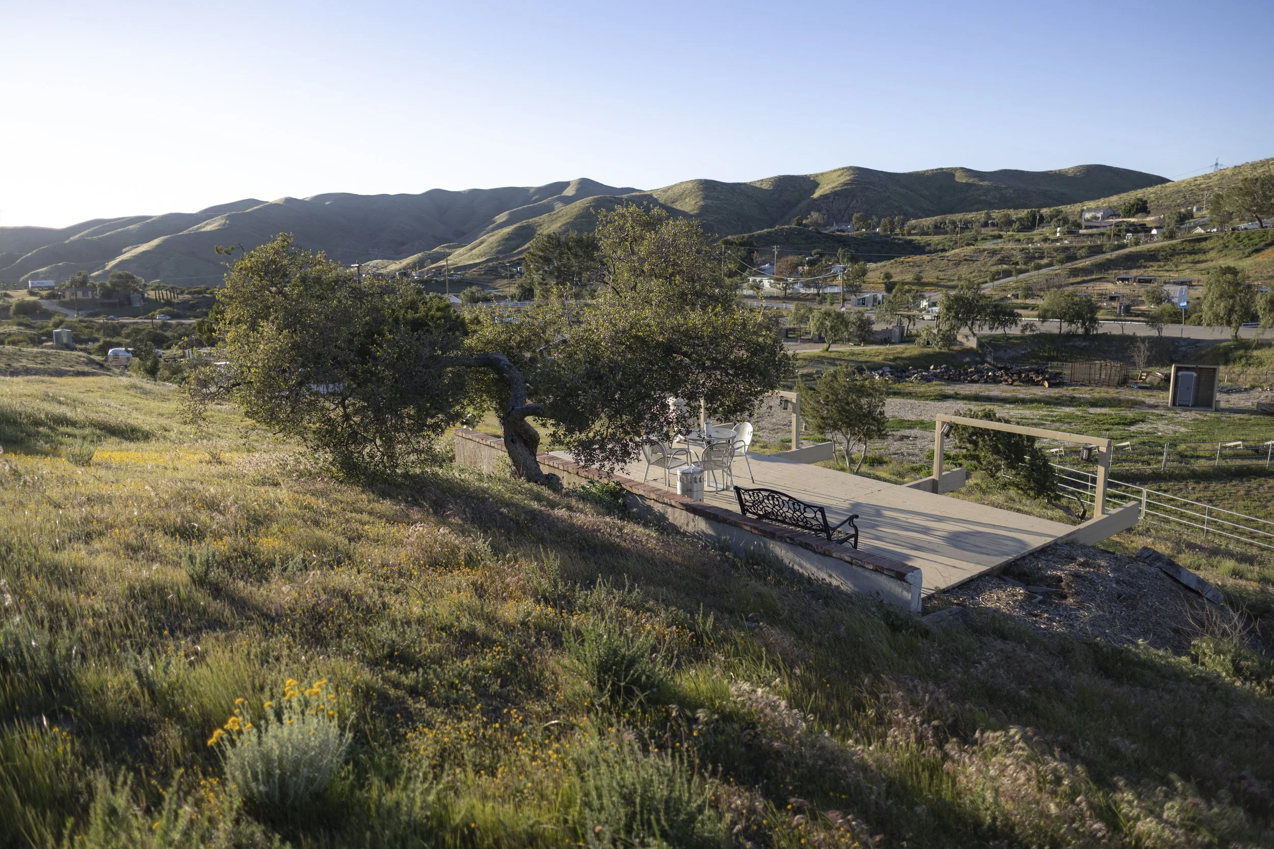 A scenic view of a hillside with a wooden deck and a picnic table, surrounded by green trees and wildflowers, with hills and a small town in the background under a clear sky.