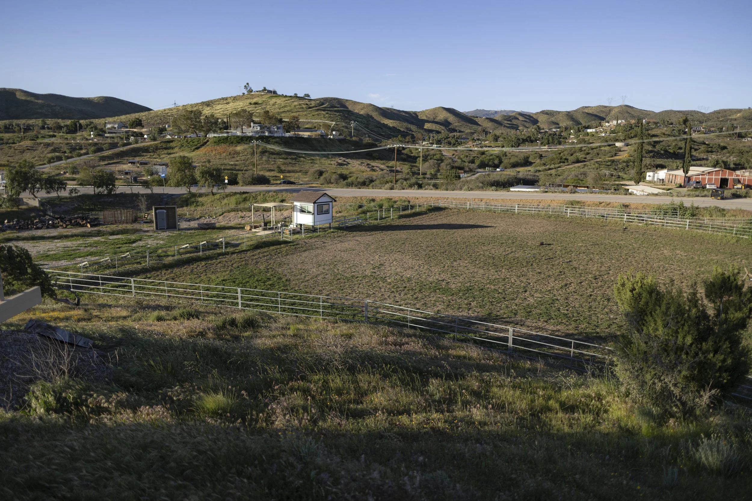 Countryside landscape with rolling hills, a fenced field, small buildings, and sparse trees under a clear blue sky.
