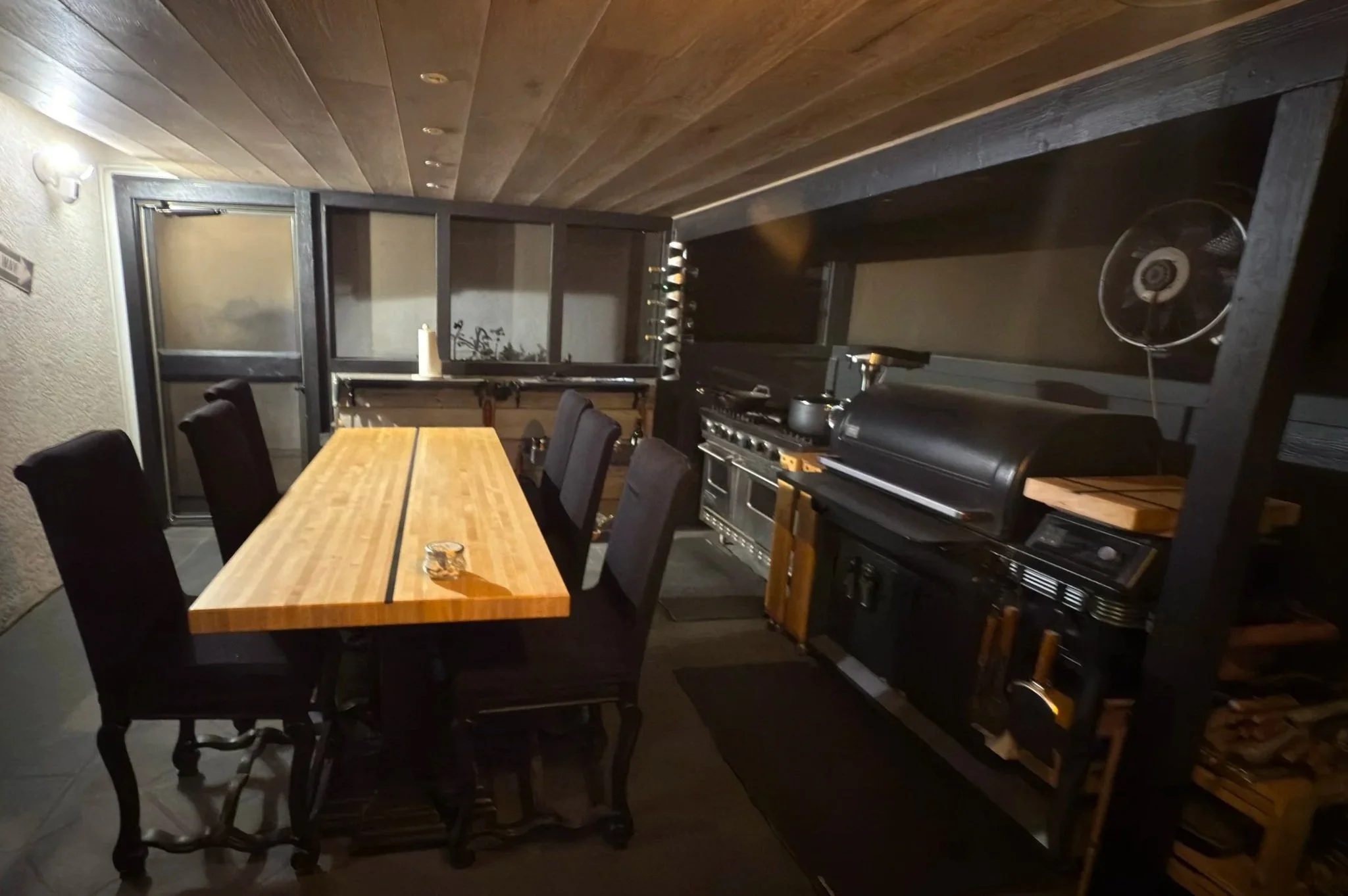 Outdoor kitchen with a wooden dining table and six chairs, grill, and cooking appliances under a wooden ceiling with lighting.
