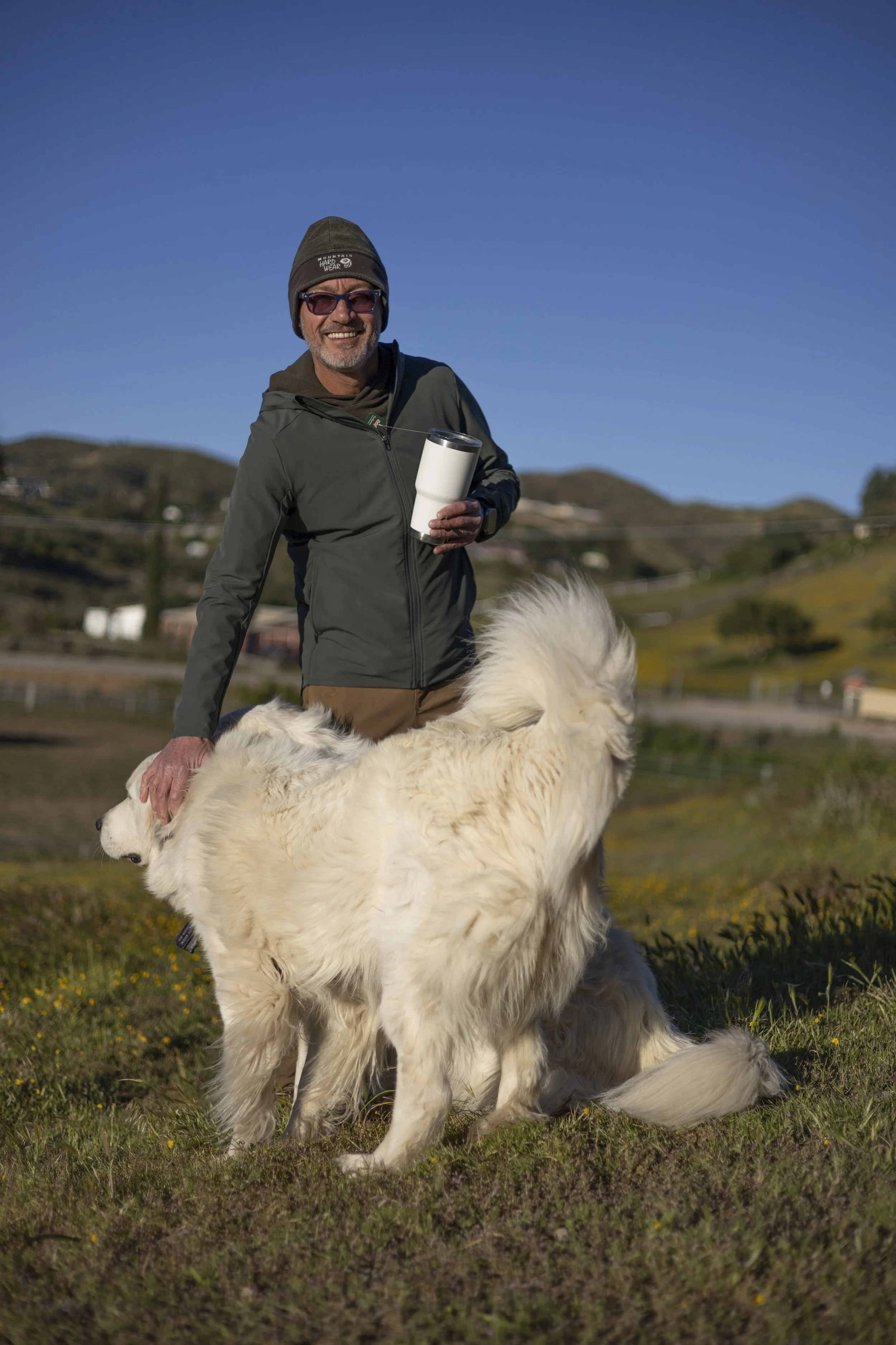 A man smiling at the camera, wearing a beanie, sunglasses, a dark jacket, and holding a white tumbler, stands outdoors on a sunny day with a large white fluffy dog sitting in front of him on a grassy field with hills and blue sky in the background.