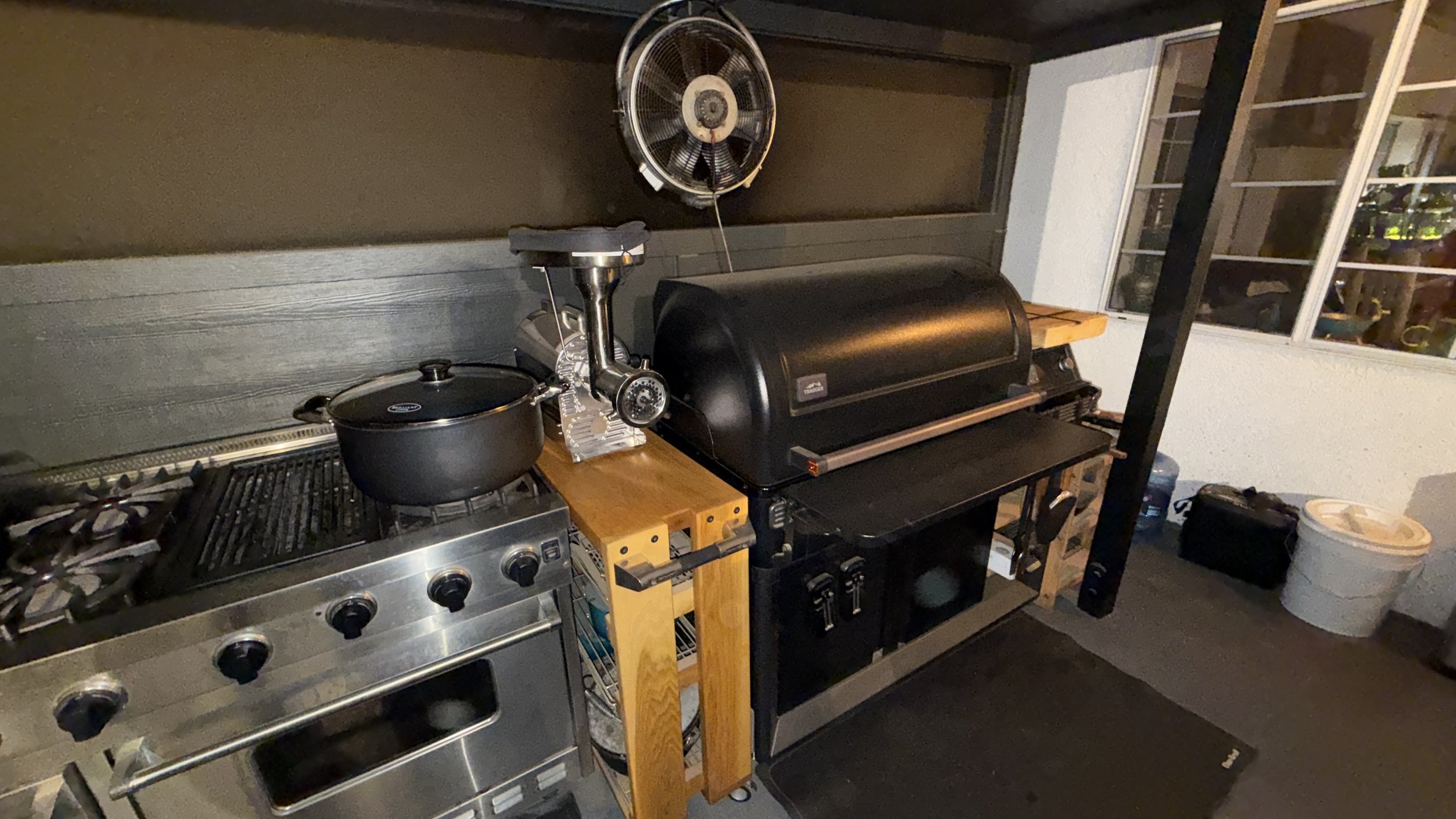 Outdoor kitchen with a gas grill, a black smoker grill, a meat grinder, a pot on the stove, a wall fan, and a window with shelves inside.