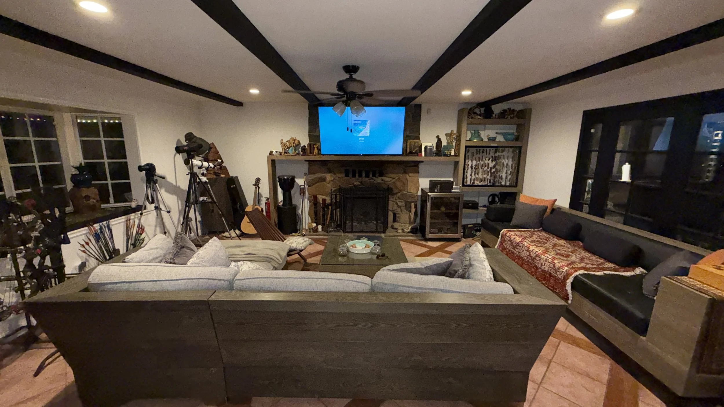 Living room with a stone fireplace, a mounted television, guitars, and telescopes. Sofas surround a coffee table with a bowl and candles. The room has wooden accents, large windows, and display shelves.