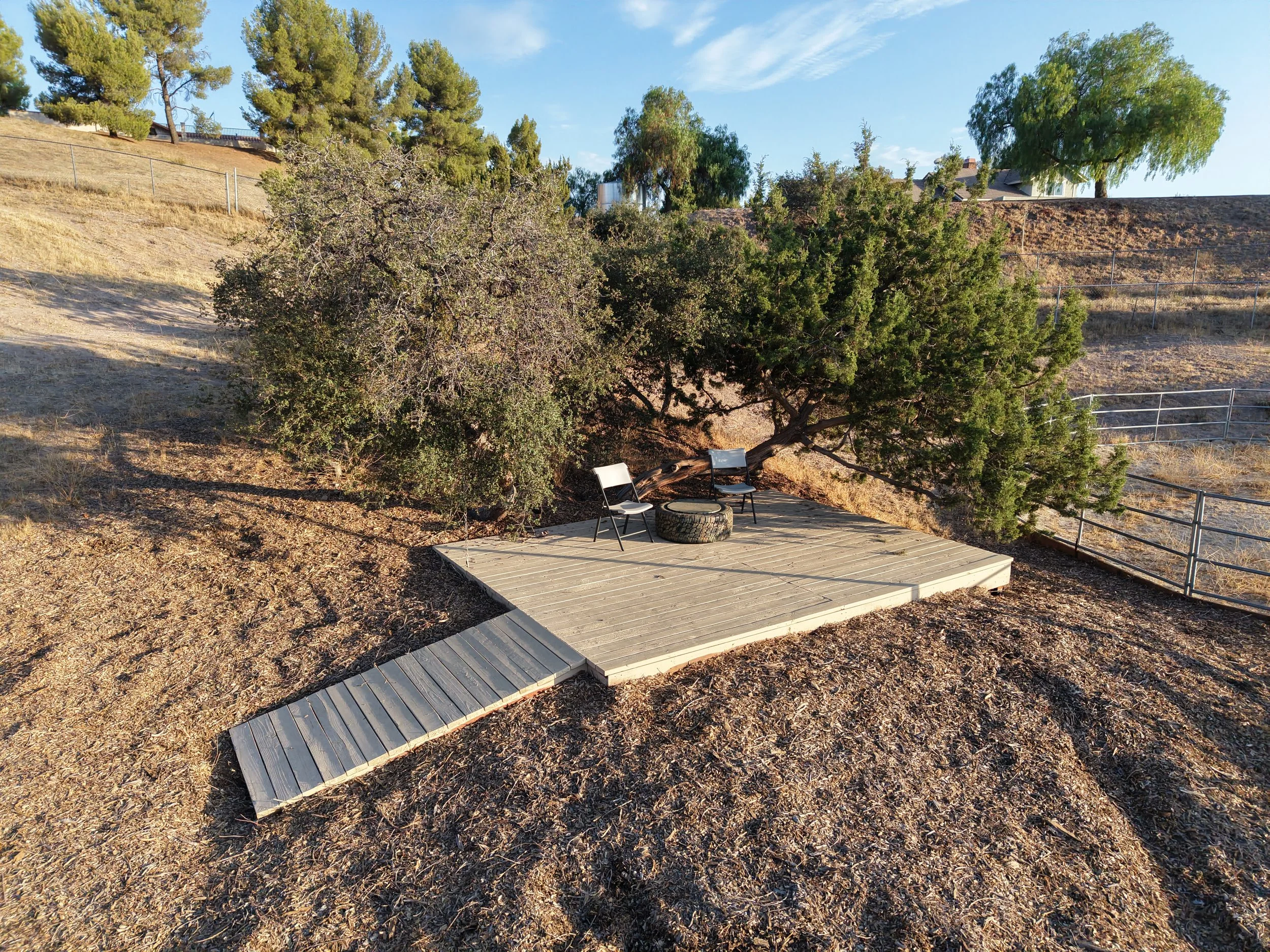 A small outdoor wooden deck with two chairs and a tire used as a table, situated under a large tree on a hillside with dry grass and a metal fence in the background.