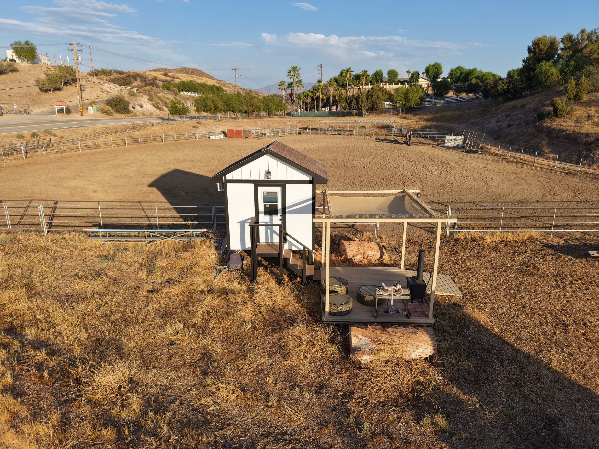 Small white shed with black trim on a fenced outdoor area on a hillside, with dry grass and trees in the background, under a partly cloudy sky.