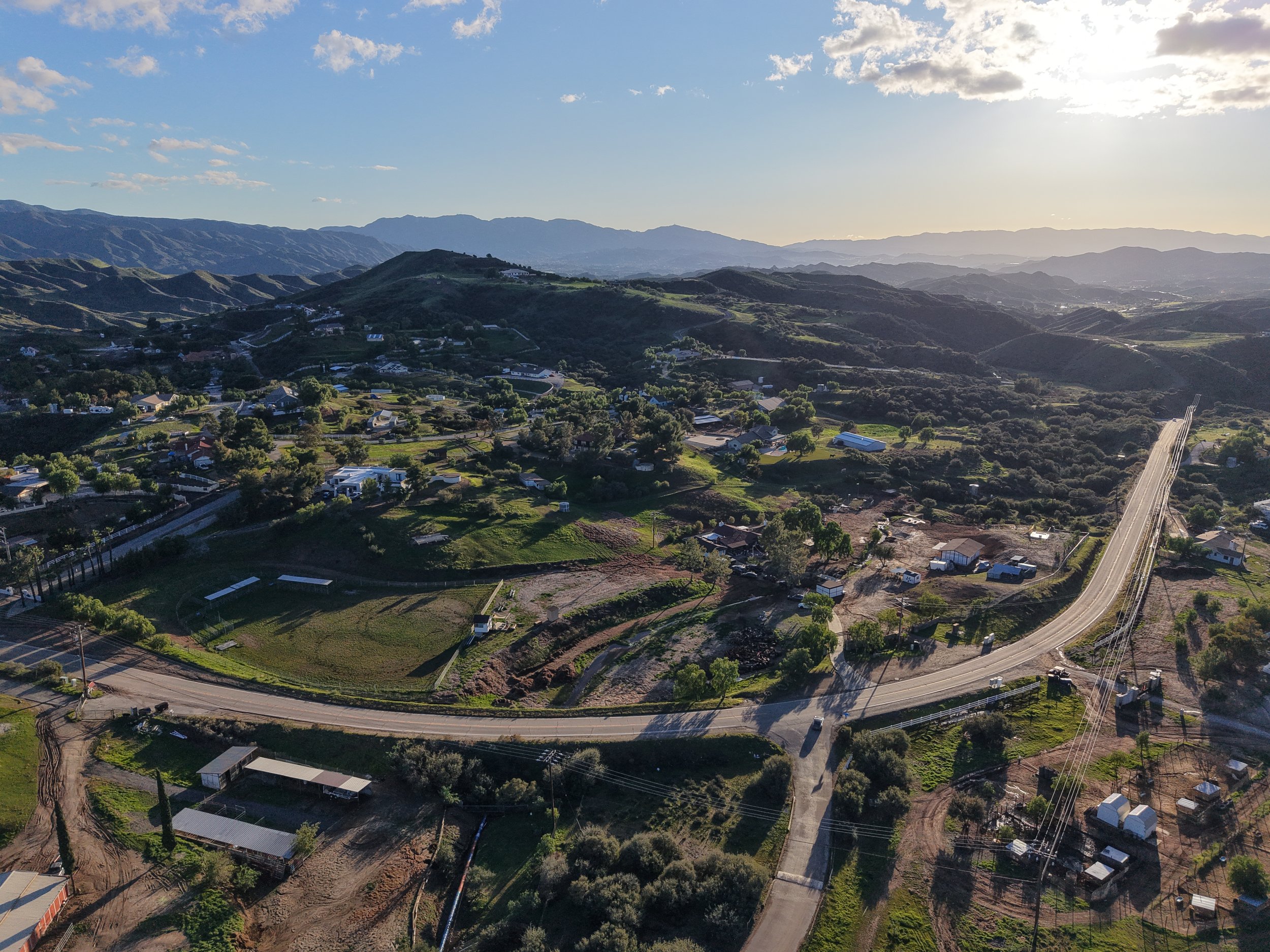 Aerial view of a rural landscape with rolling hills, scattered houses, and a winding road, taken during sunset.