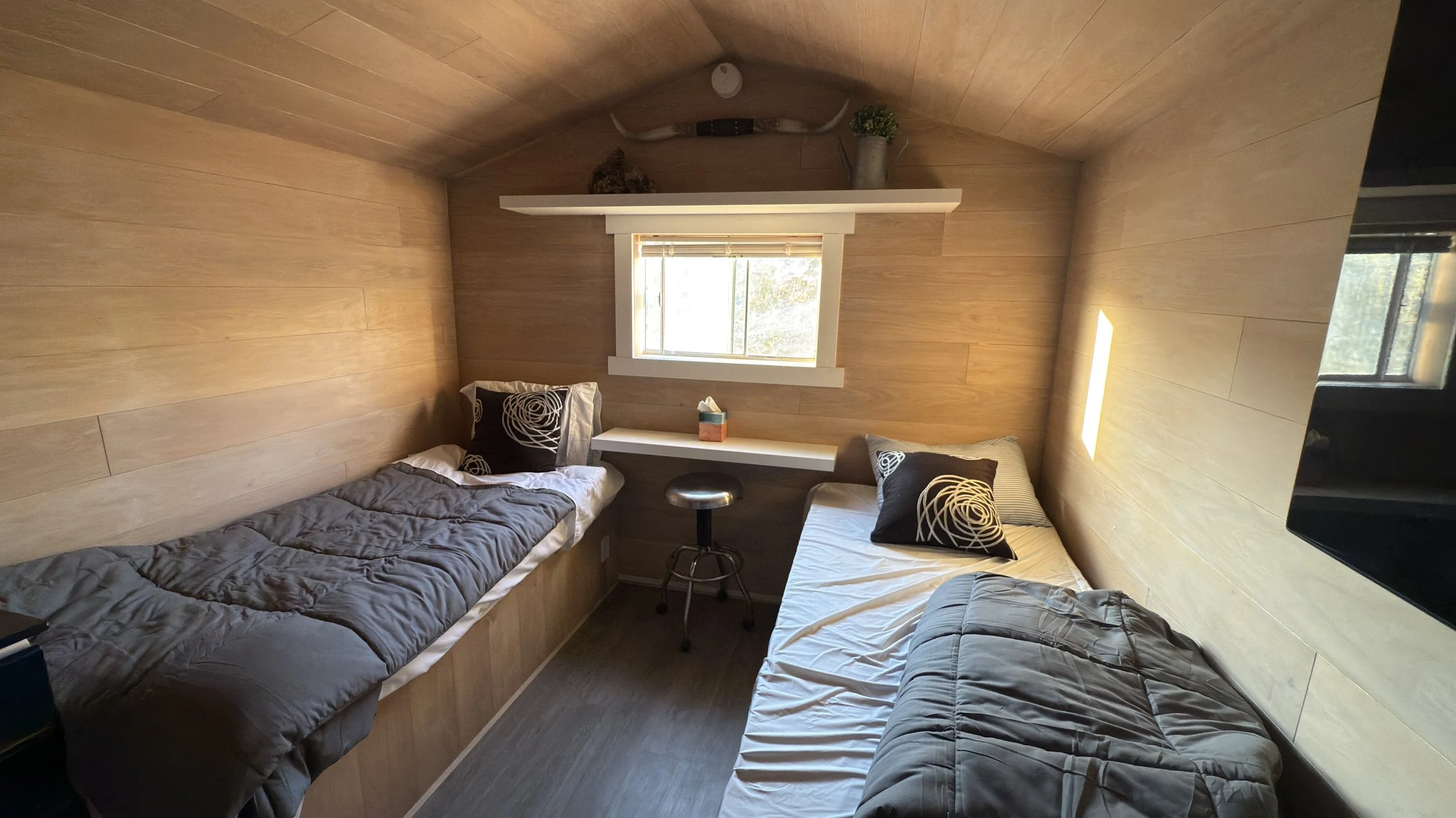 Small bedroom with two twin beds, each with black and white pillows and gray bedding, a window with blinds, a small white shelf with a tissue box, a stool, and a shelf with decorative items, all inside a wooden-paneled room.
