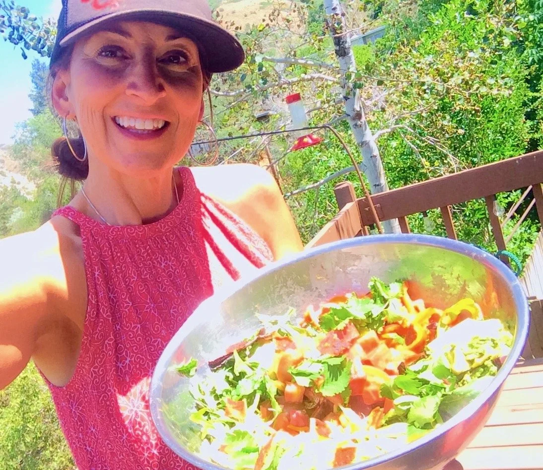 Woman smiling outdoors holding a bowl of salad with vegetables and greens, wearing a pink sleeveless top and a gray cap.