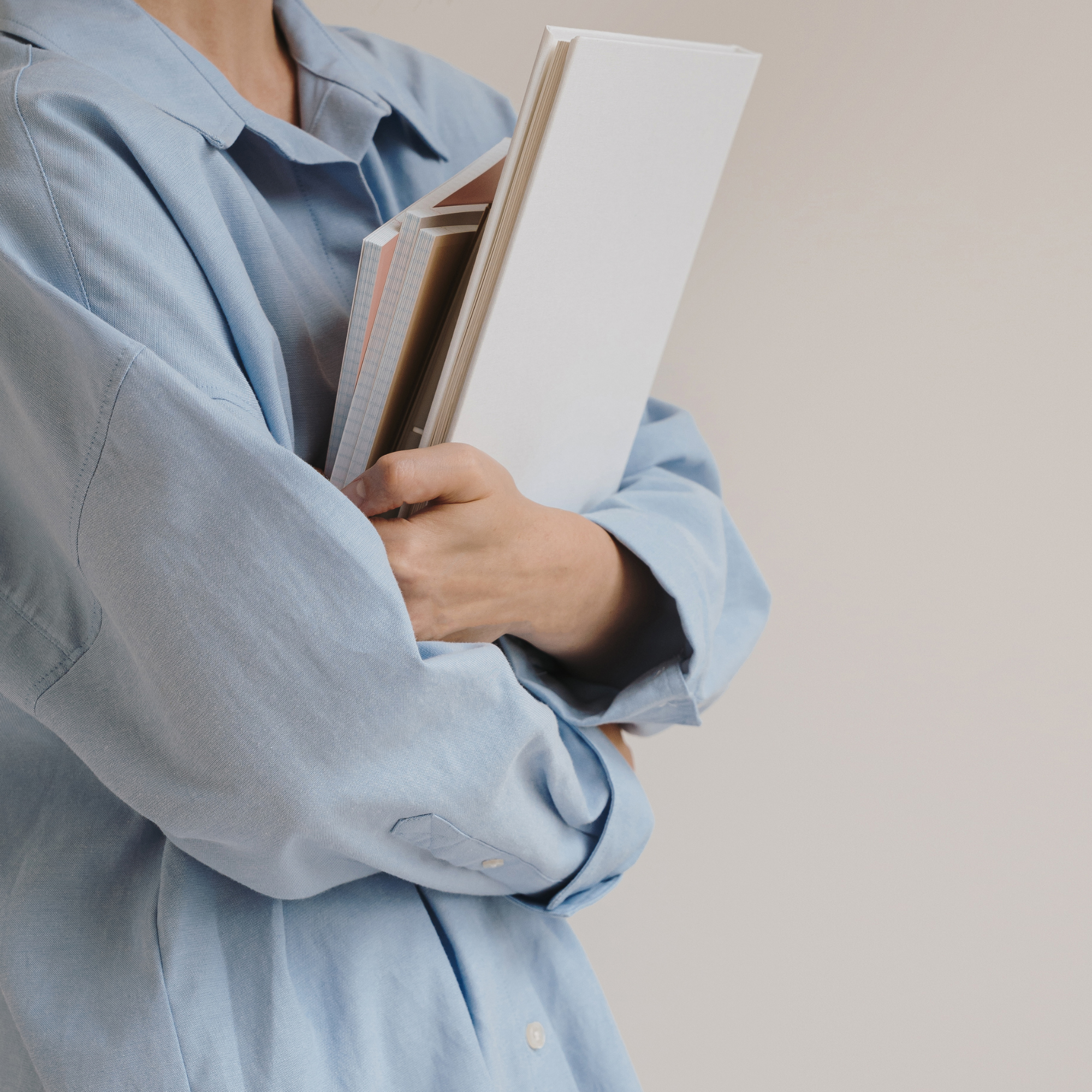 Person in a light blue shirt holding a stack of notebooks and folders against a plain wall.