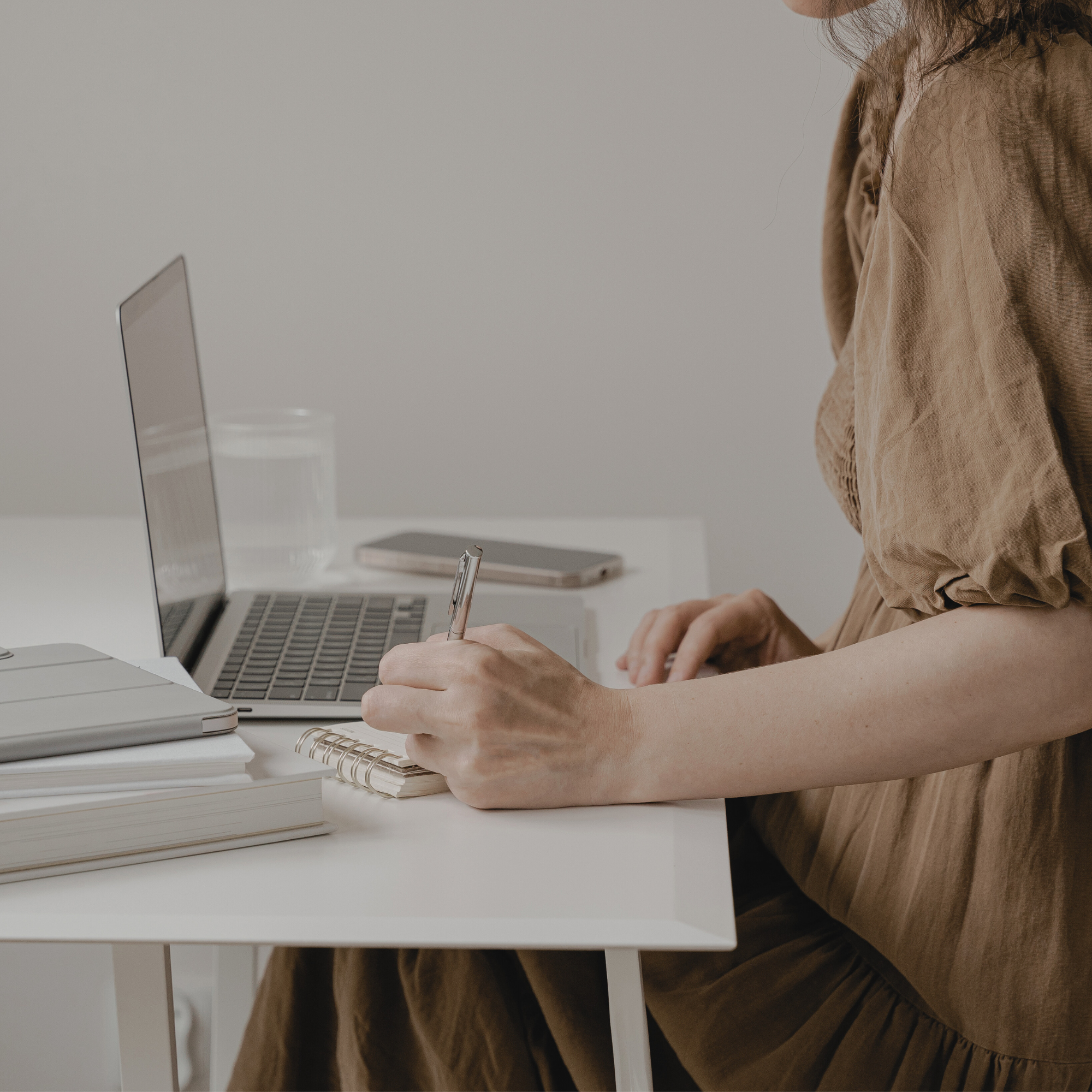 A woman sitting at a white desk working with a laptop, taking notes in a spiral notebook, with a smartphone and a glass of water on the desk.
