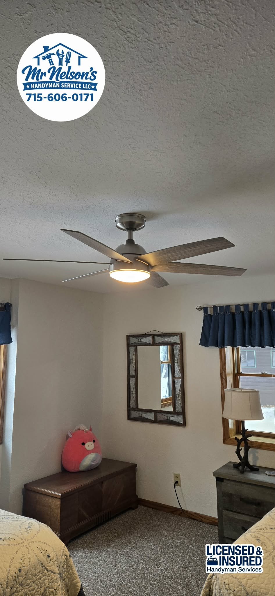A bedroom with a ceiling fan, a wall mirror, a window with blue curtains, a lamp on a gray nightstand, and a pink plush pig toy on a wooden storage bench.