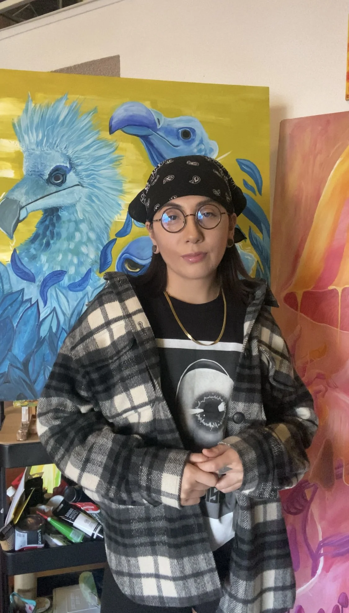 Contemporary Artist Cassandra Carrillo, in a black bandana, and plaid jacket standing in front of colorful animal paintings, including a blue bird and a mythological surreal paintings, in art studio.
