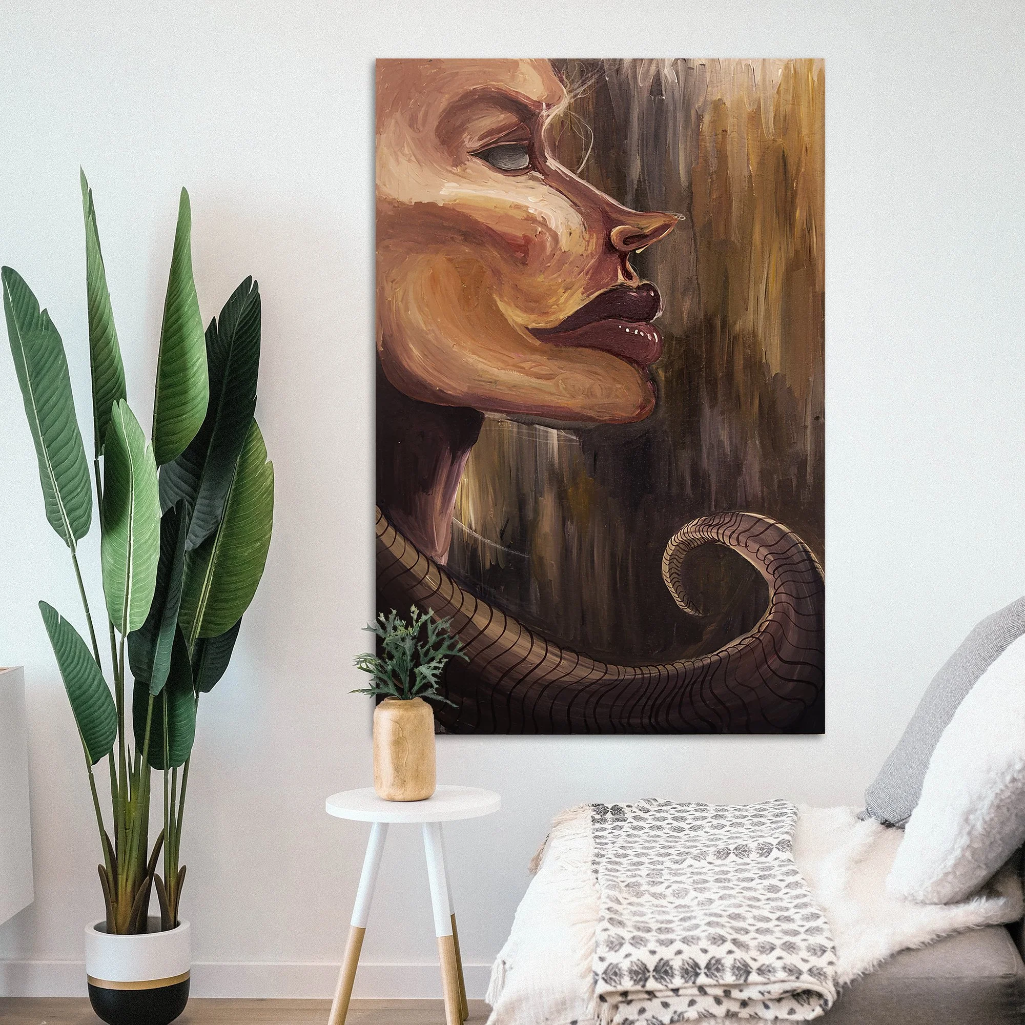 Interior of a modern living room with a large abstract portrait of a woman's face on the wall, a tall potted plant with large green leaves, and a small round white side table with a beige vase and green plant. There's a gray couch with throw pillows and a patterned blanket.