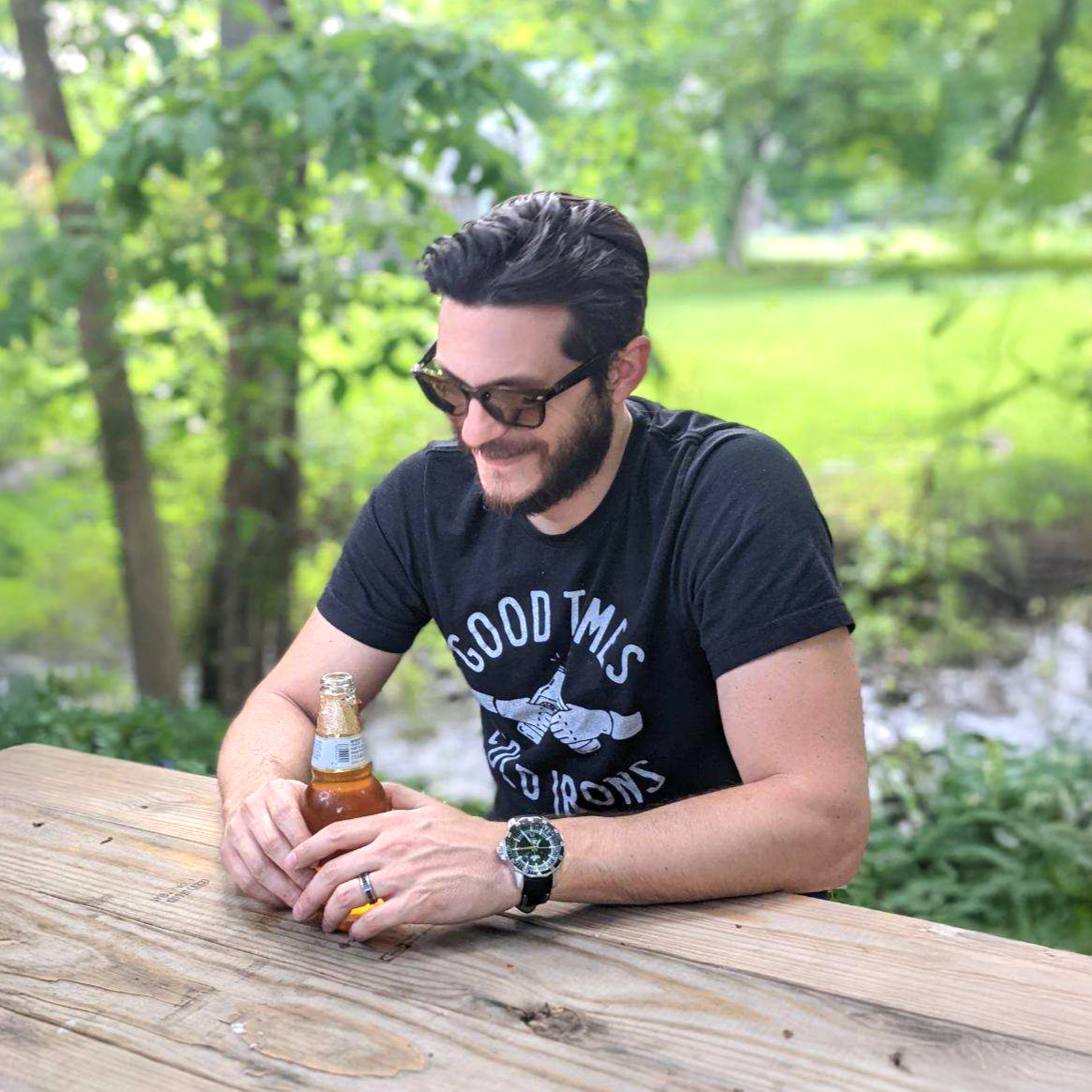 Jeremie Musyt, Pittsburgh creative director, sitting outdoors at a wooden table wearing sunglasses and holding a beer.