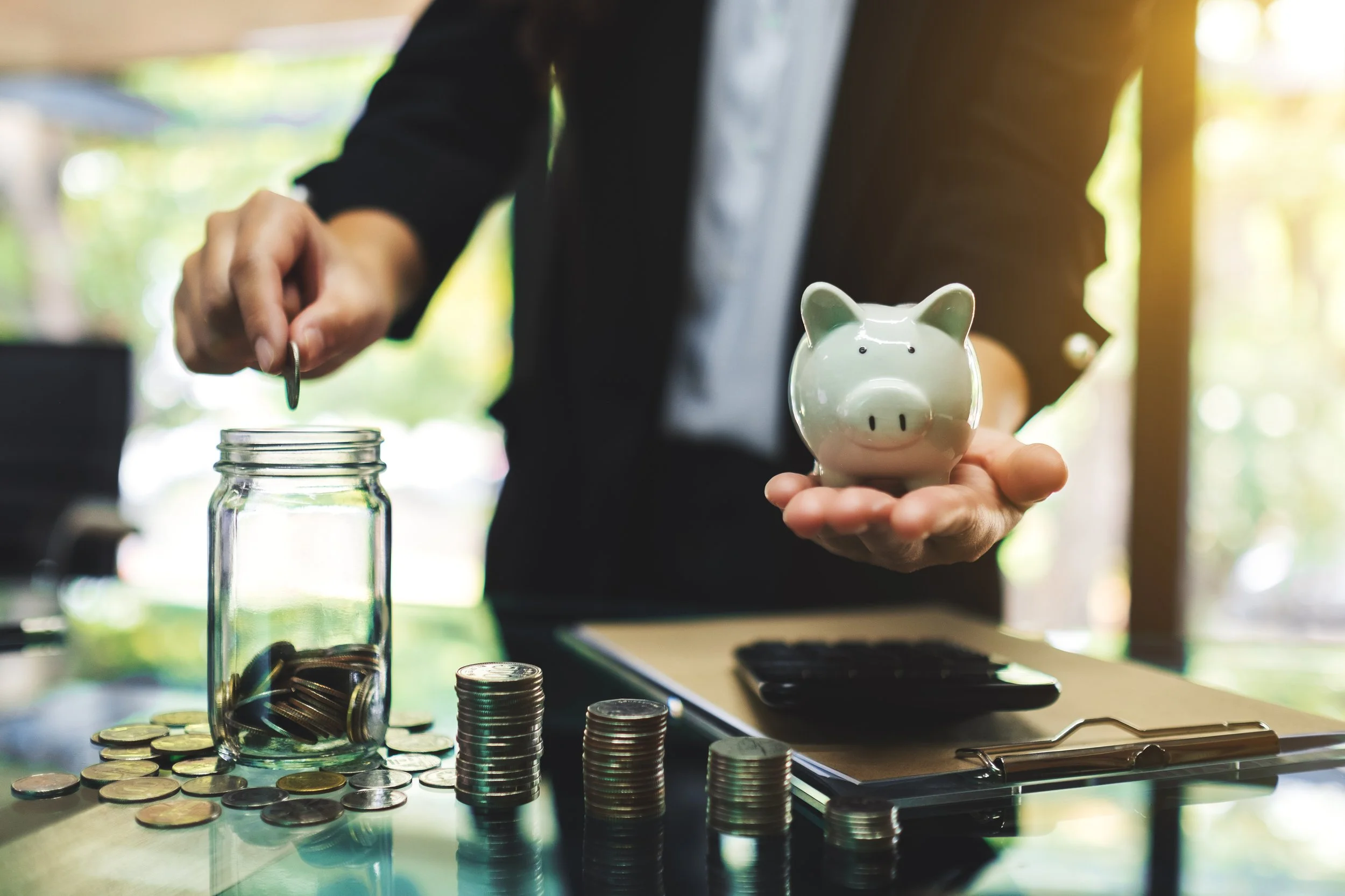 Una persona sosteniendo una alcancía con forma de cerdito y colocando una moneda en ella, con monedas y una taza de vidrio con monedas dentro sobre una mesa.