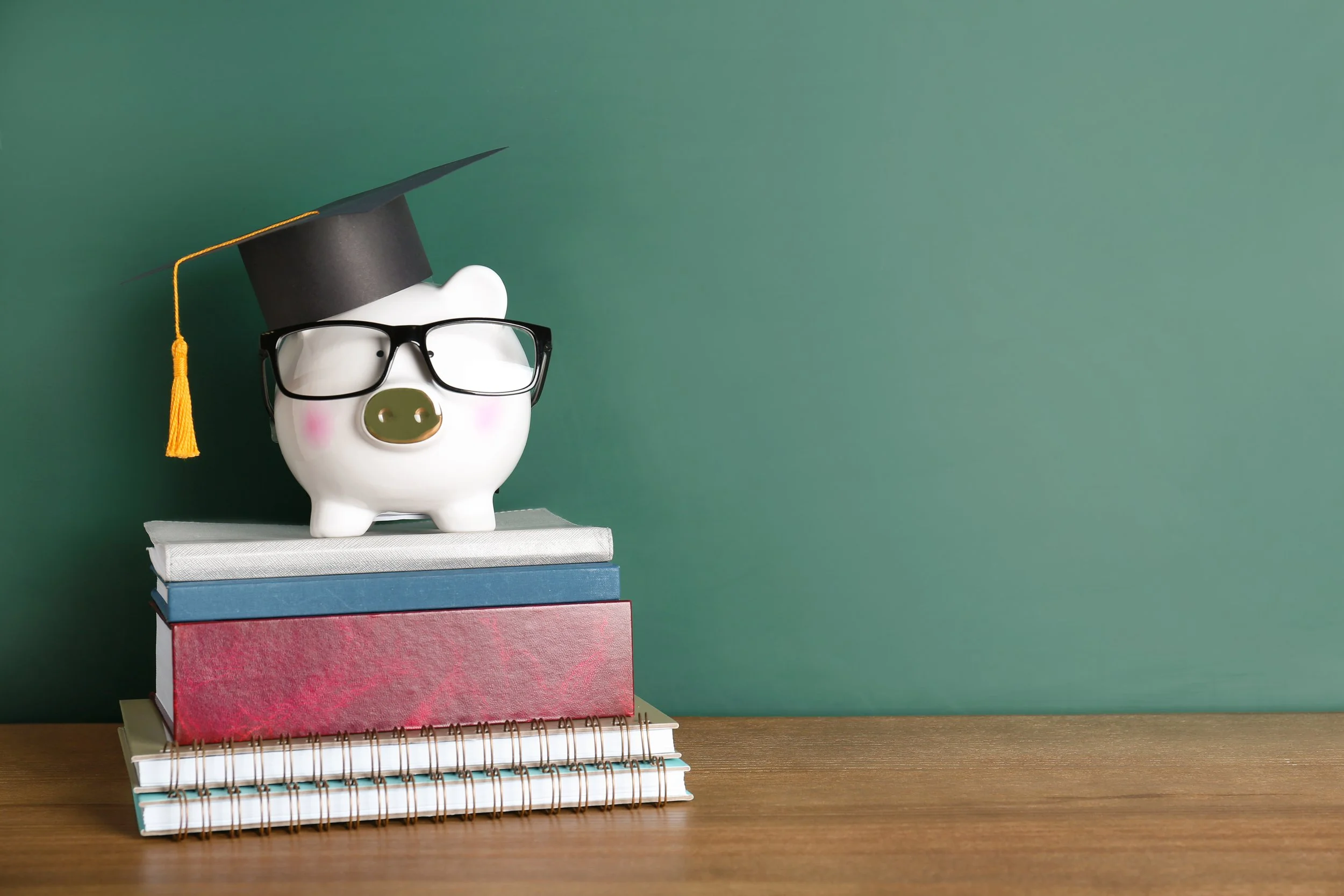Cerdito de cerámica con gafas y sombrero de graduación, sobre pila de libros en escritorio de madera, fondo color verde.