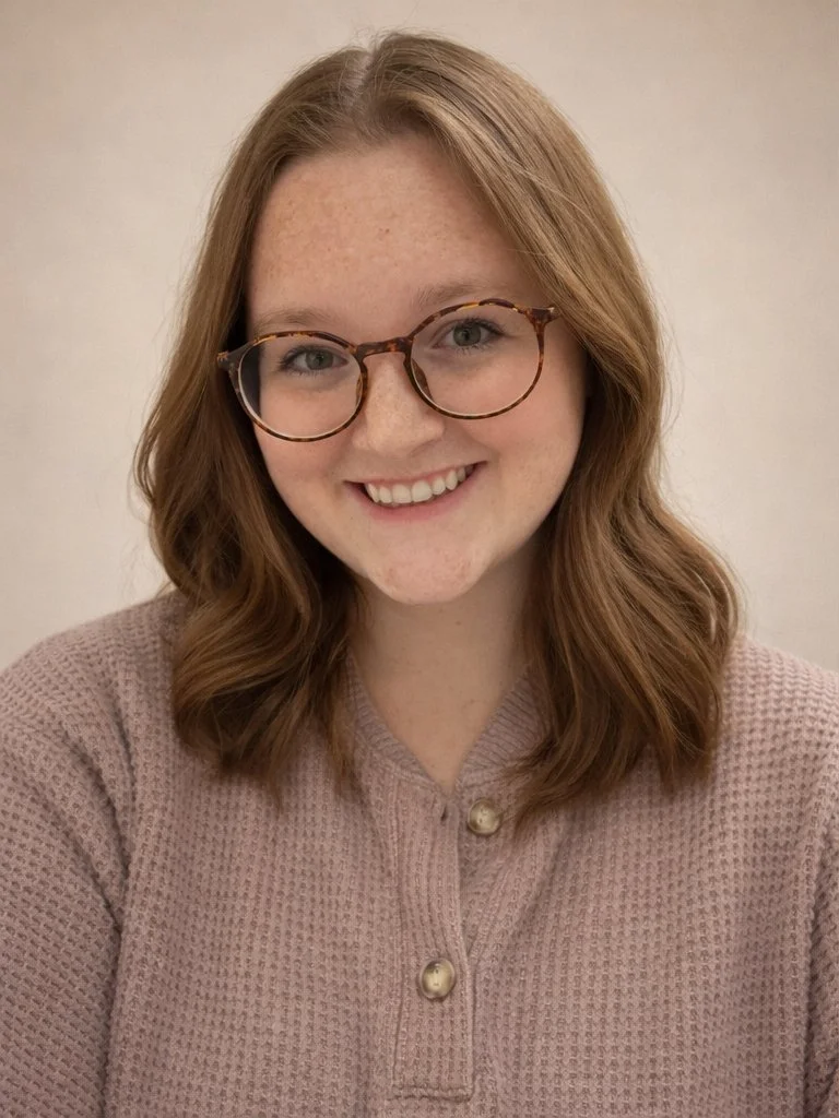 A young woman with wavy red hair, glasses, and a beige textured sweater smiling at the camera against a plain light-colored background.