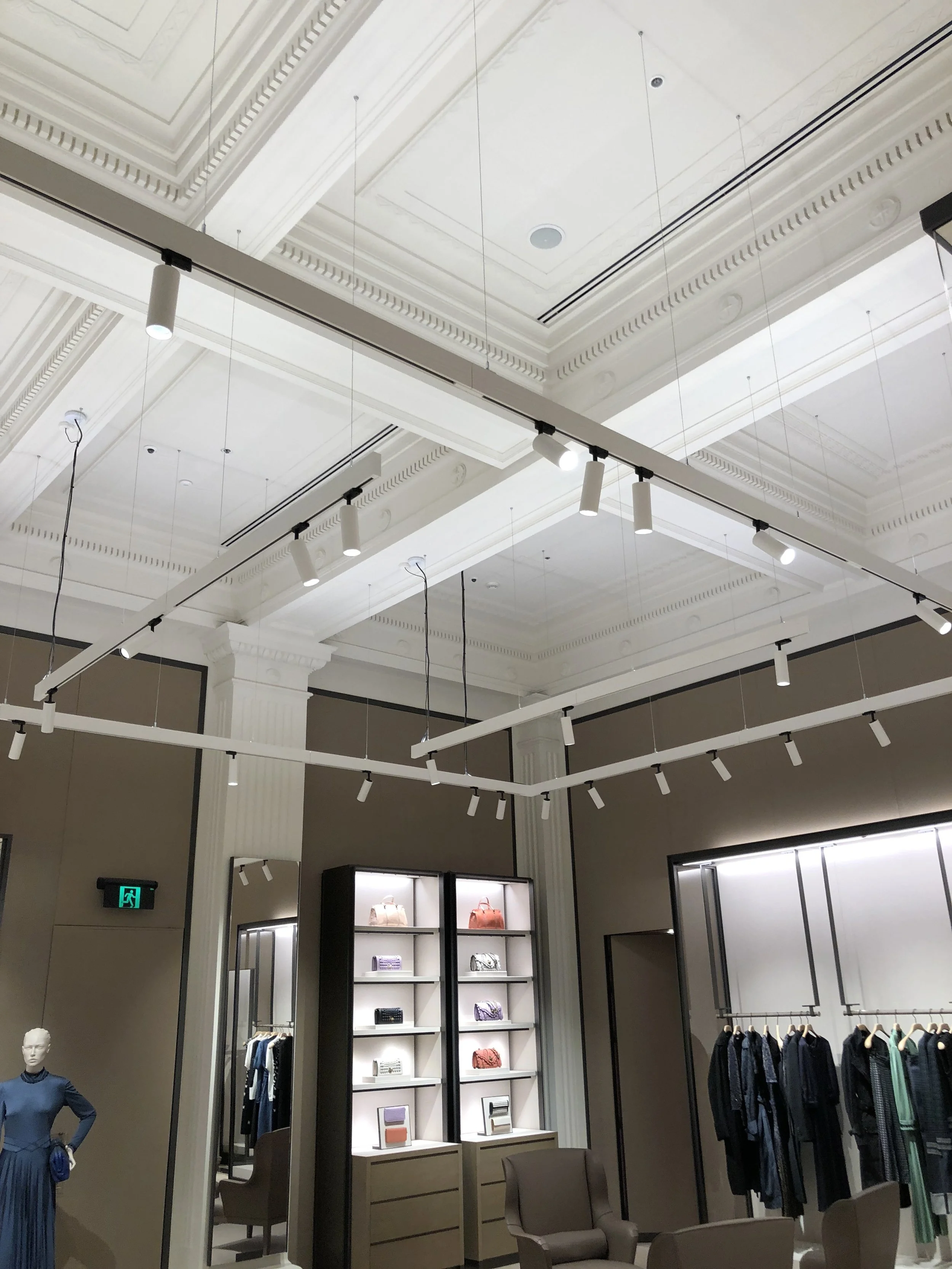 Interior of a luxury retail store with clothing racks, display shelves with handbags, mannequins, and modern lighting fixtures on high ceilings.