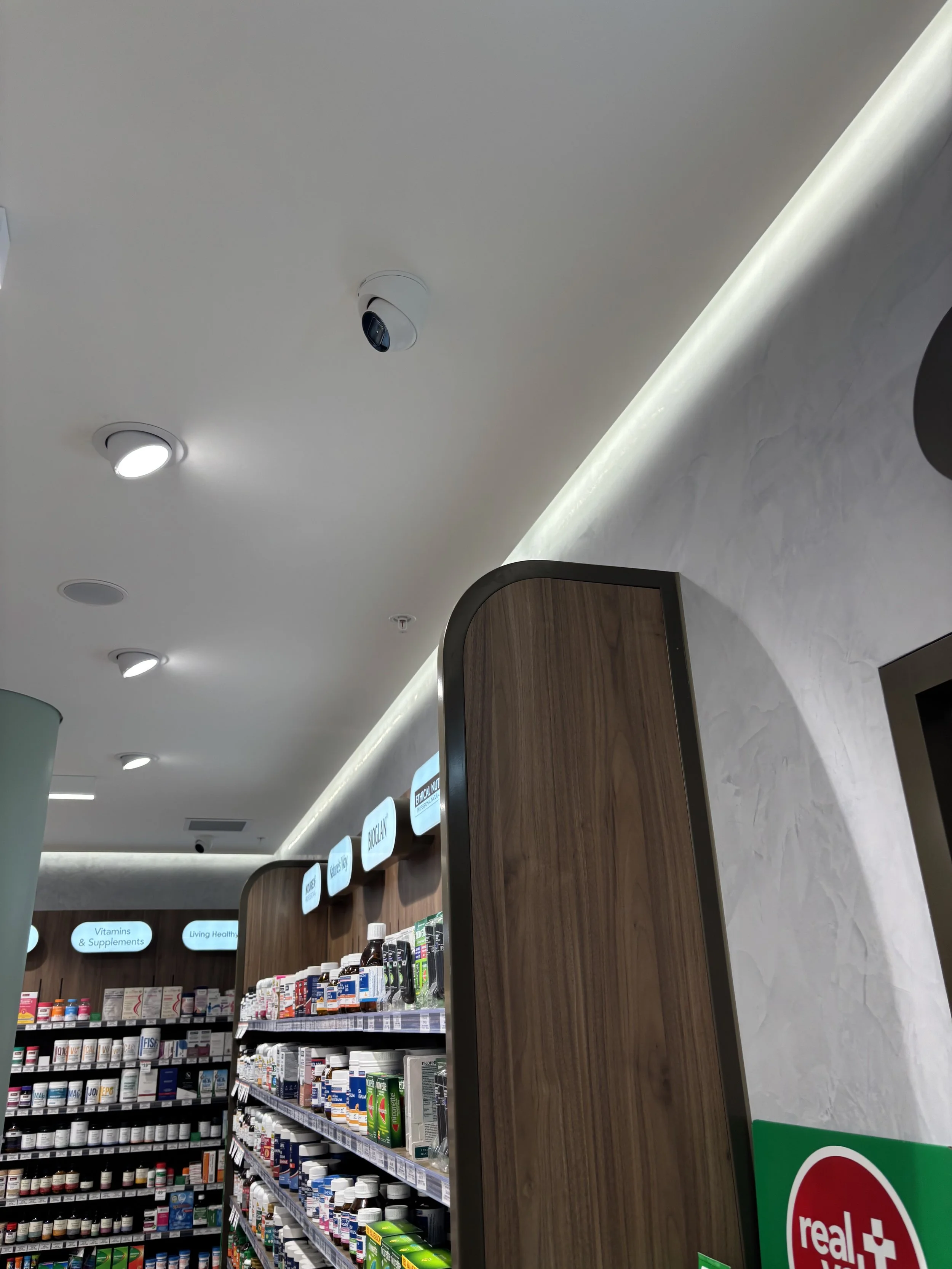 Inside a pharmacy or health store with shelves stocked with vitamins, supplements, and medicines, and ceiling lights and security cameras visible.
