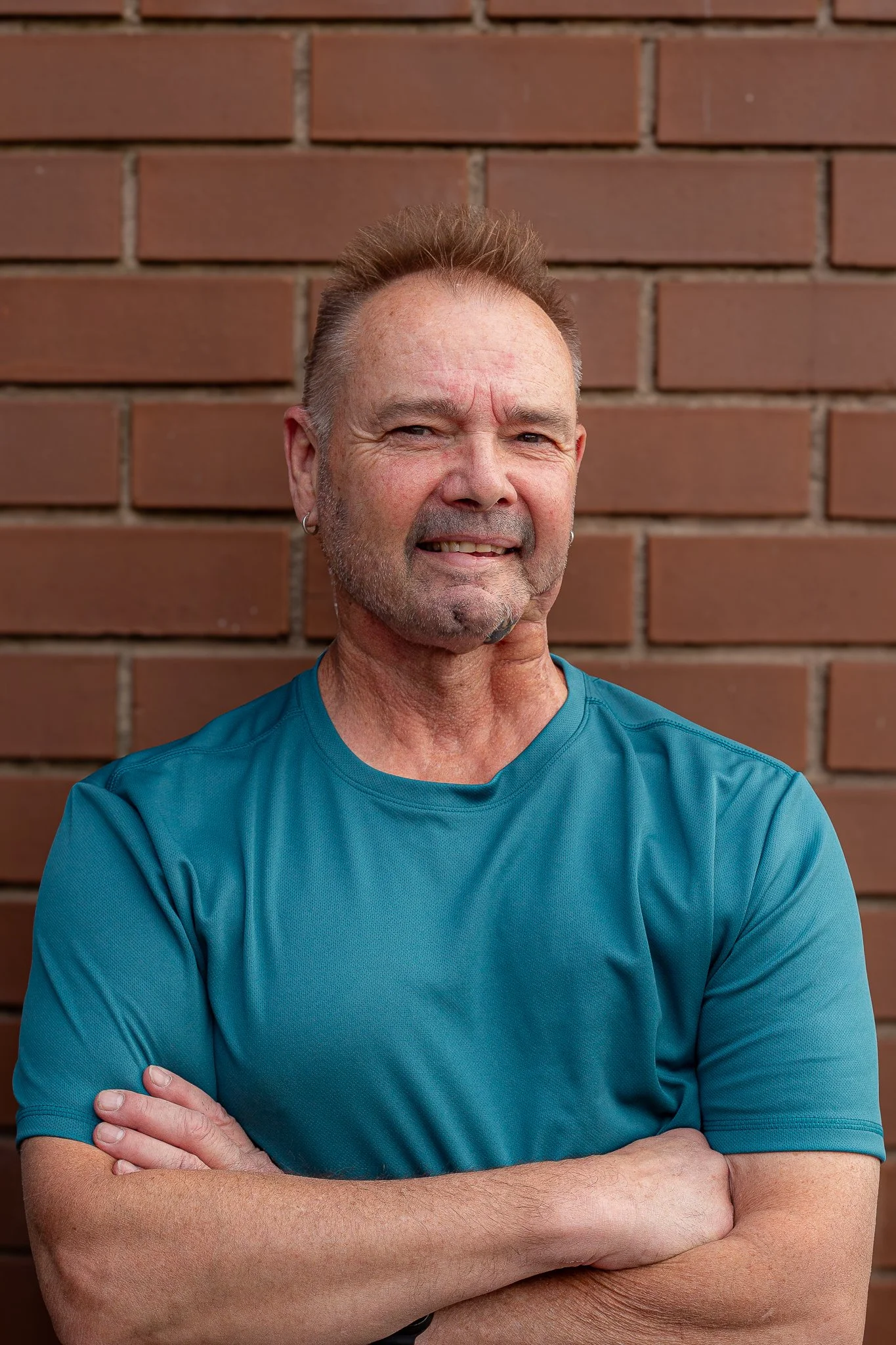 A middle-aged man with short, spiked hair, wearing a teal t-shirt, stands with his arms crossed in front of a red brick wall, looking at the camera with a slight smile.