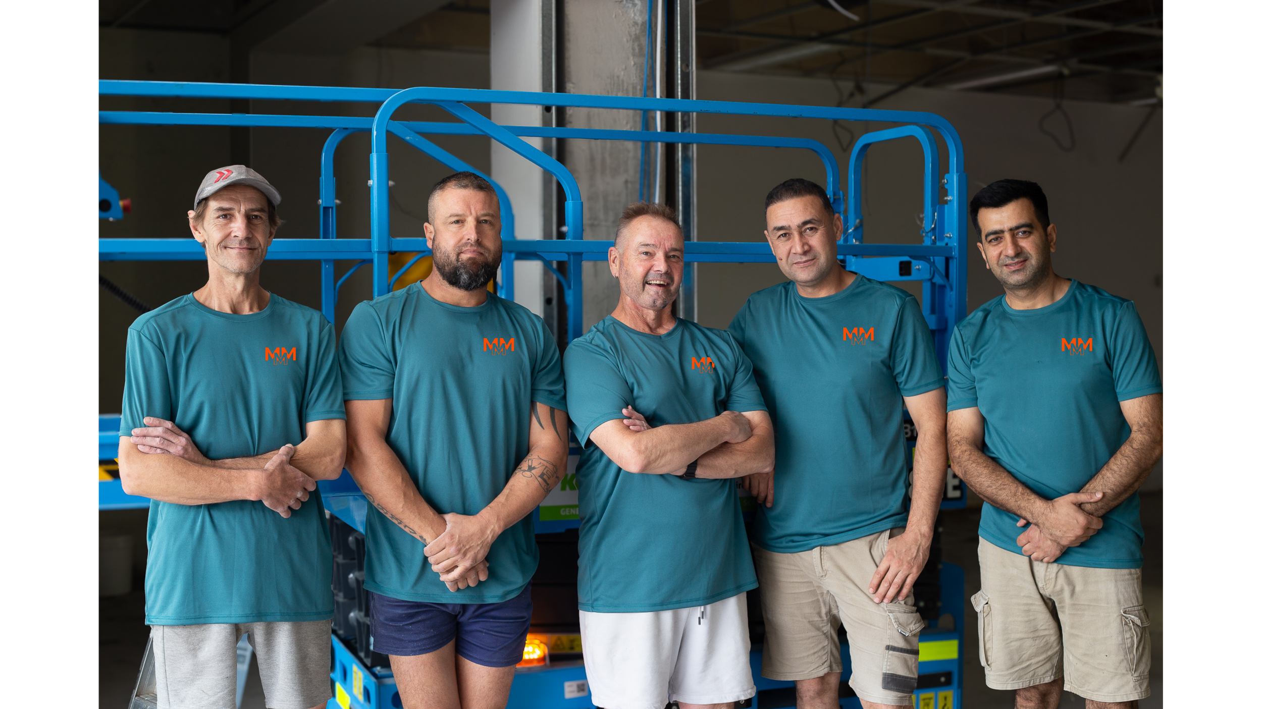 Five men standing in front of a blue scissor lift, all wearing matching teal shirts with an orange logo, inside a warehouse or industrial space.