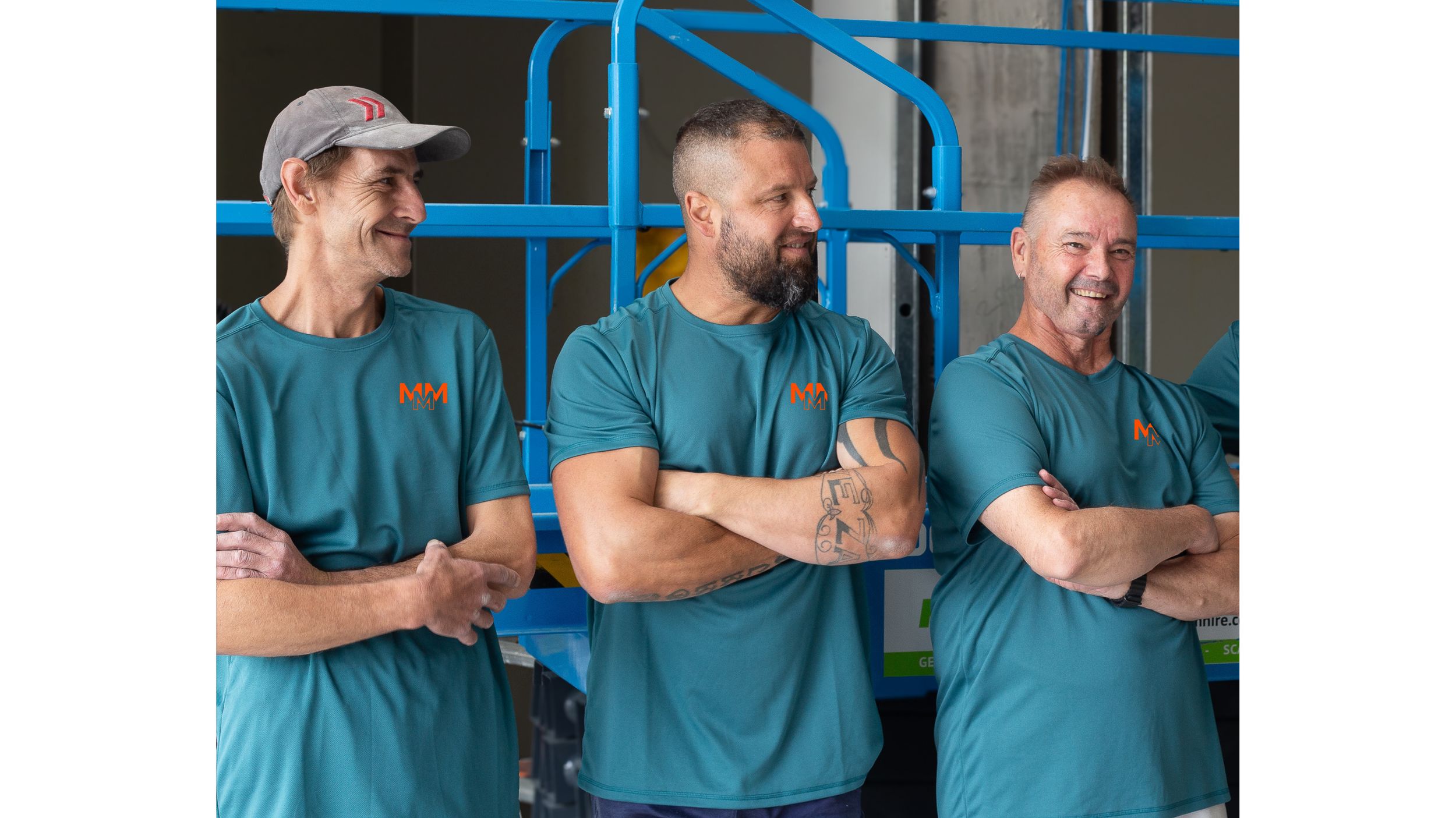 Three men standing with arms crossed, wearing teal shirts with a red and black logo, smiling, in an industrial setting with blue equipment and concrete walls.