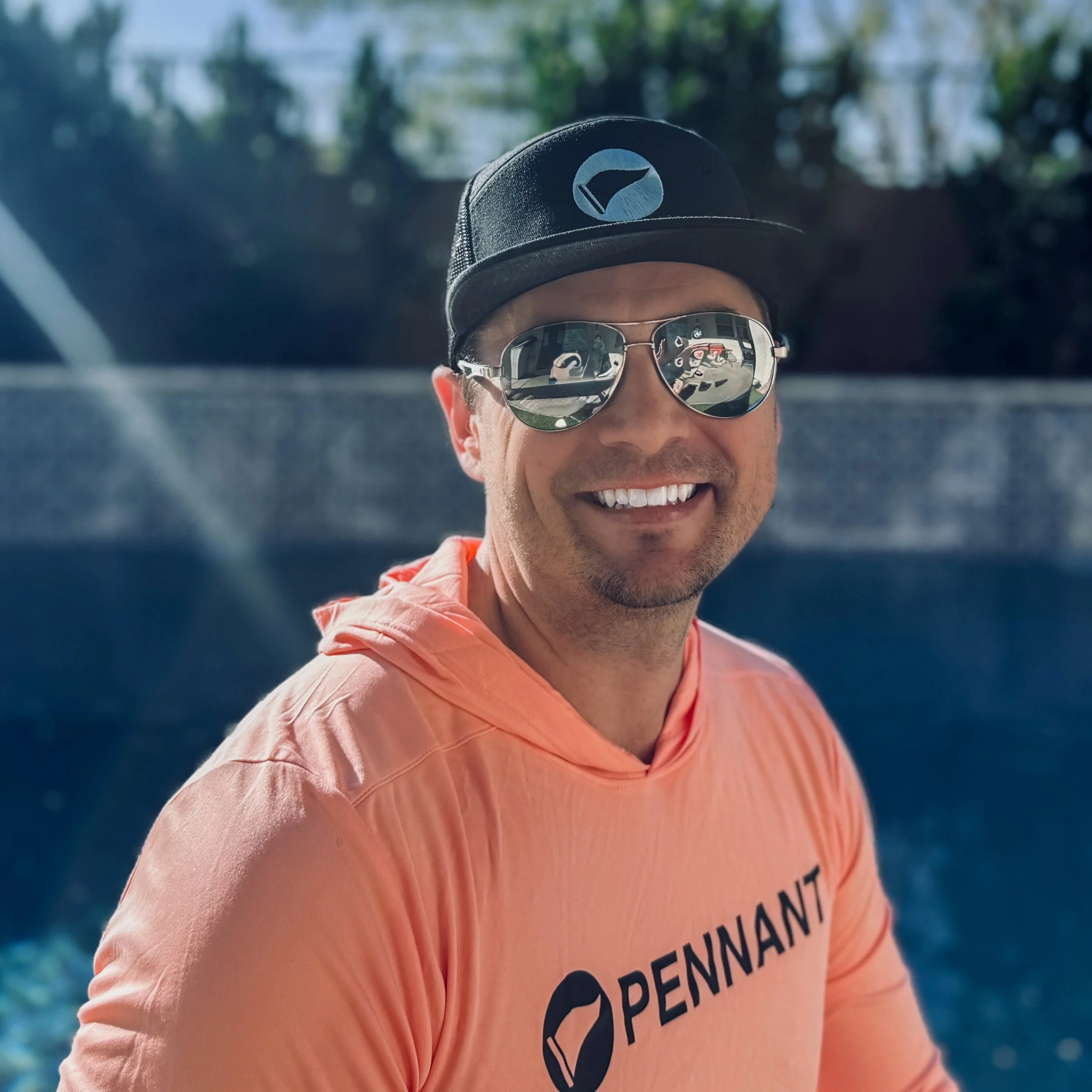 Smiling man wearing sunglasses, a black cap with a Pennant logo, and an orange Pennant hoodie, outdoors near a swimming pool.