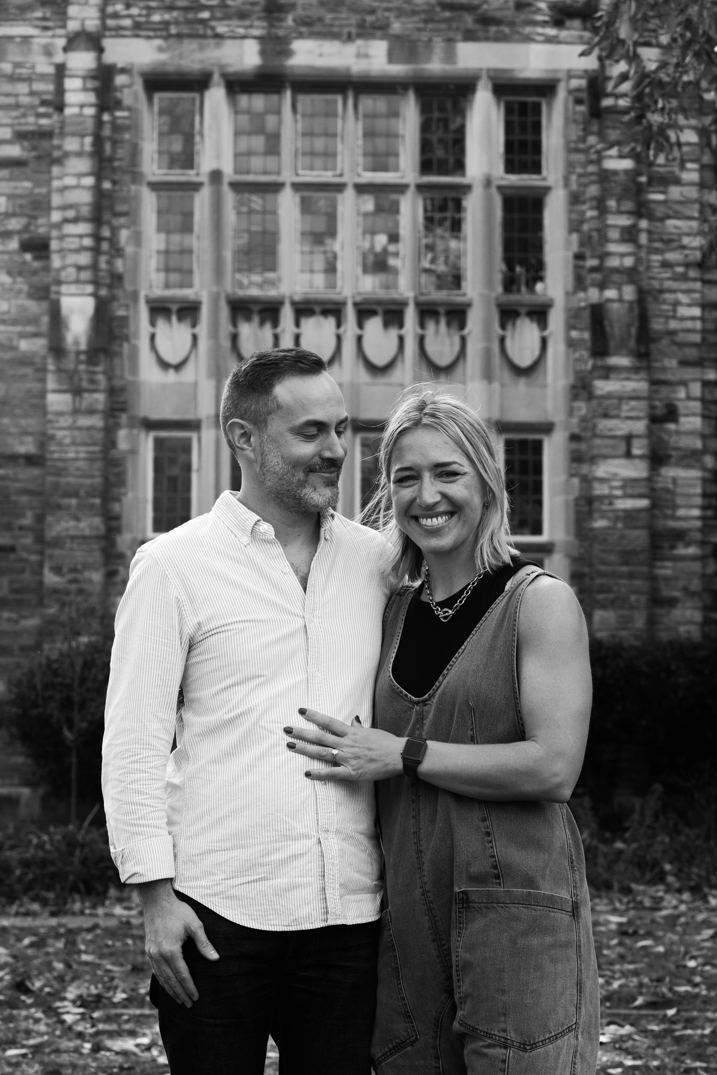 A smiling woman with blonde hair and a man with short hair and a beard standing in front of a historic building with large windows, outdoors. The woman is wearing a sleeveless denim dress, a black shirt, a necklace, and a watch, while the man is dressed in a light-colored striped shirt and dark pants.