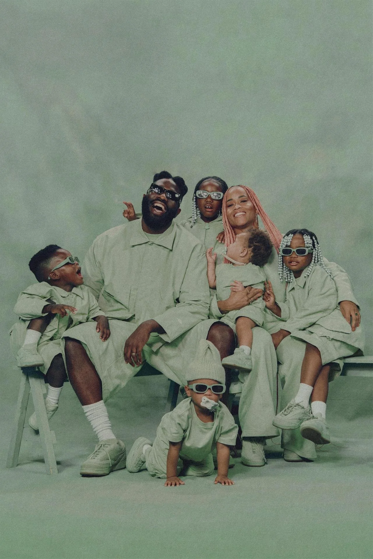 A joyful Black family of seven wearing matching light green outfits, poses in front of a plain green backdrop. The parents are seated on a bench with their children around them, some sitting on their laps and others standing beside them, all wearing 