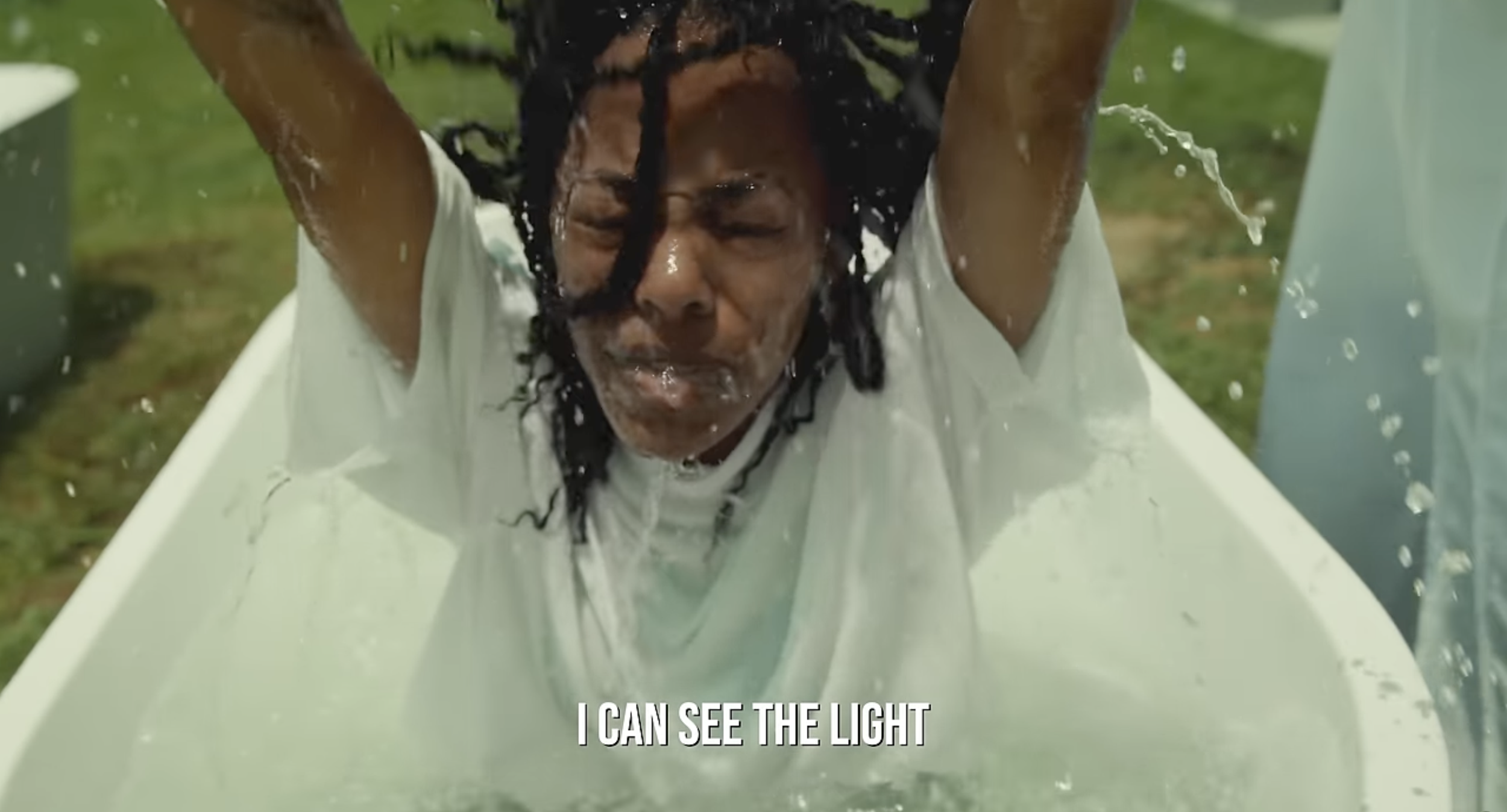 Person with dreadlocks leaning back in a white bathtub outdoors, eyes closed, with water splashing around, wearing a white shirt, with the caption 'I CAN SEE THE LIGHT' at the bottom.