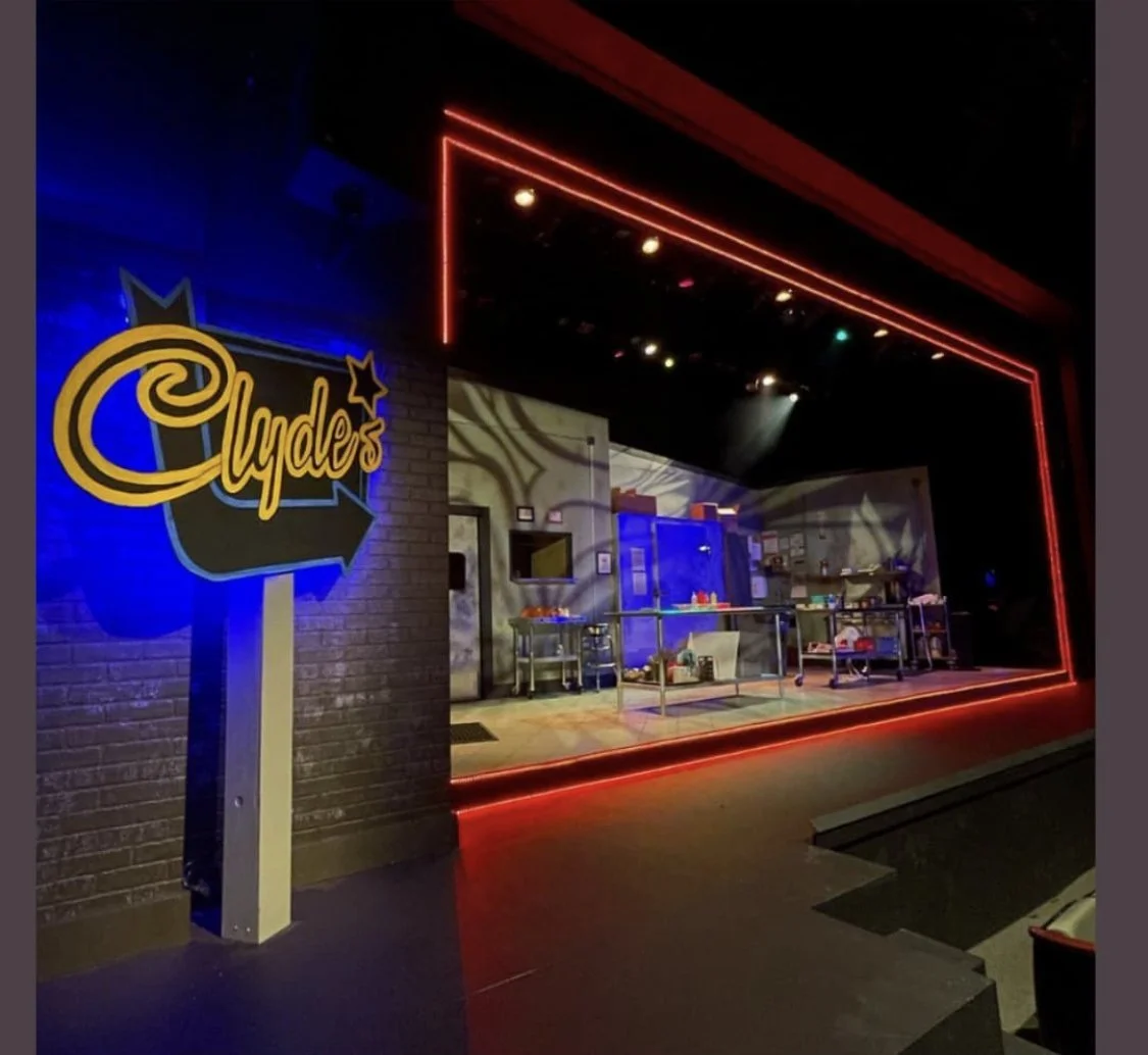 The stage of a theater with a neon sign reading "Clyde's" outside, featuring a brick wall and stage lights, set up with tables and props, ready for a performance.
