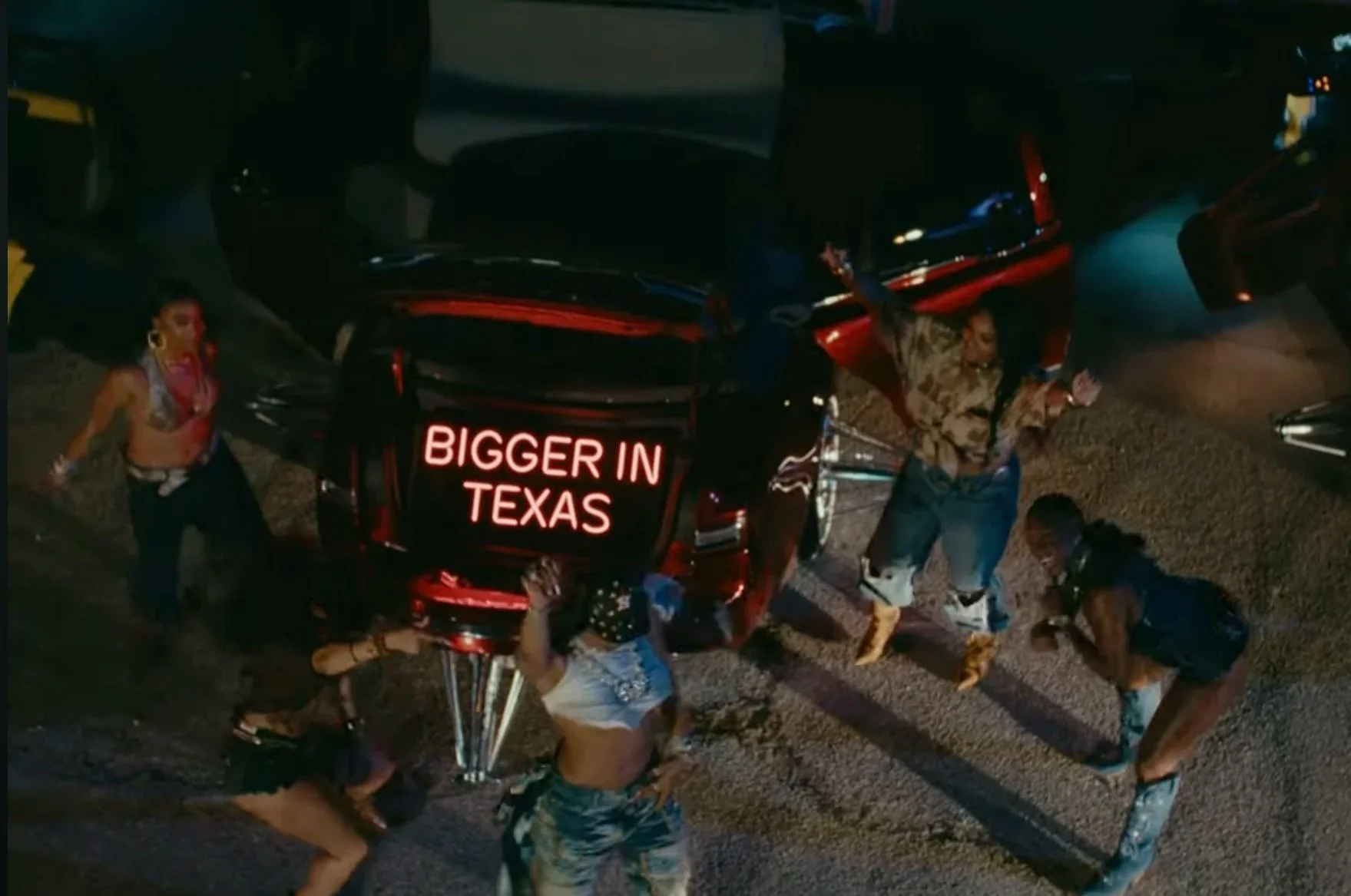 People dancing and celebrating around a vehicle with a neon sign that says 'BIGGER IN TEXAS' at night.
