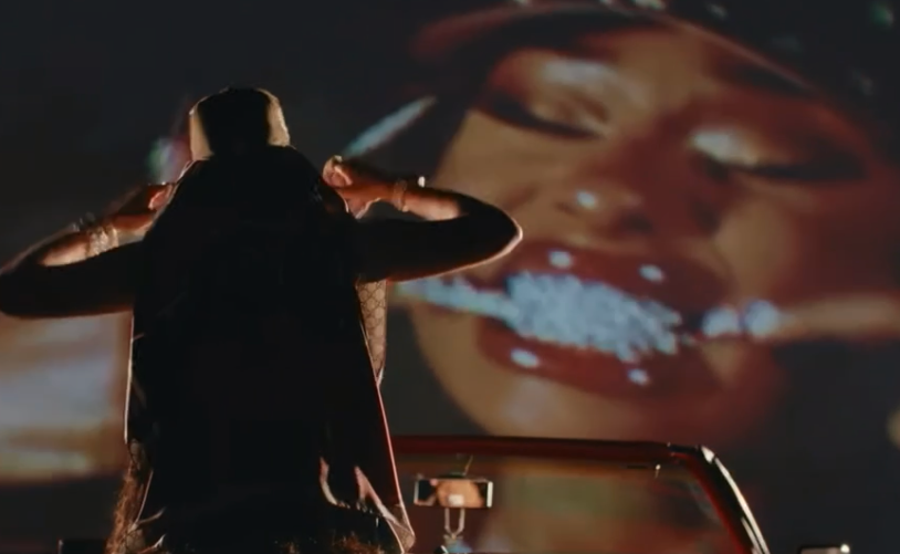 A person standing in front of a large screen displaying a close-up of a woman's face with open mouth and jewelry in her hand