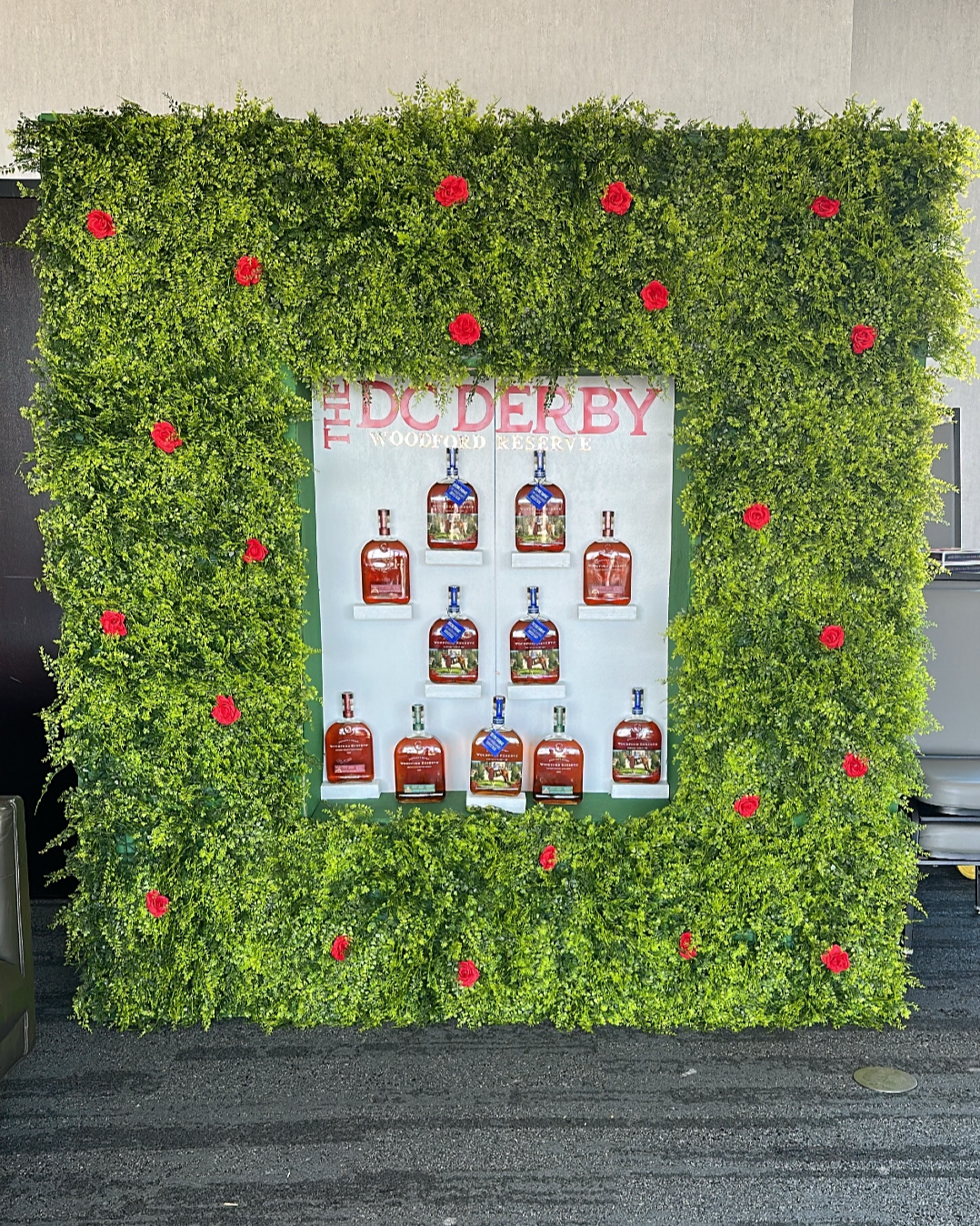 Display of Woodford Reserve bourbon bottles on a white background, framed by green bush wall with red flowers, with 'The DC Derby' sign at the top.