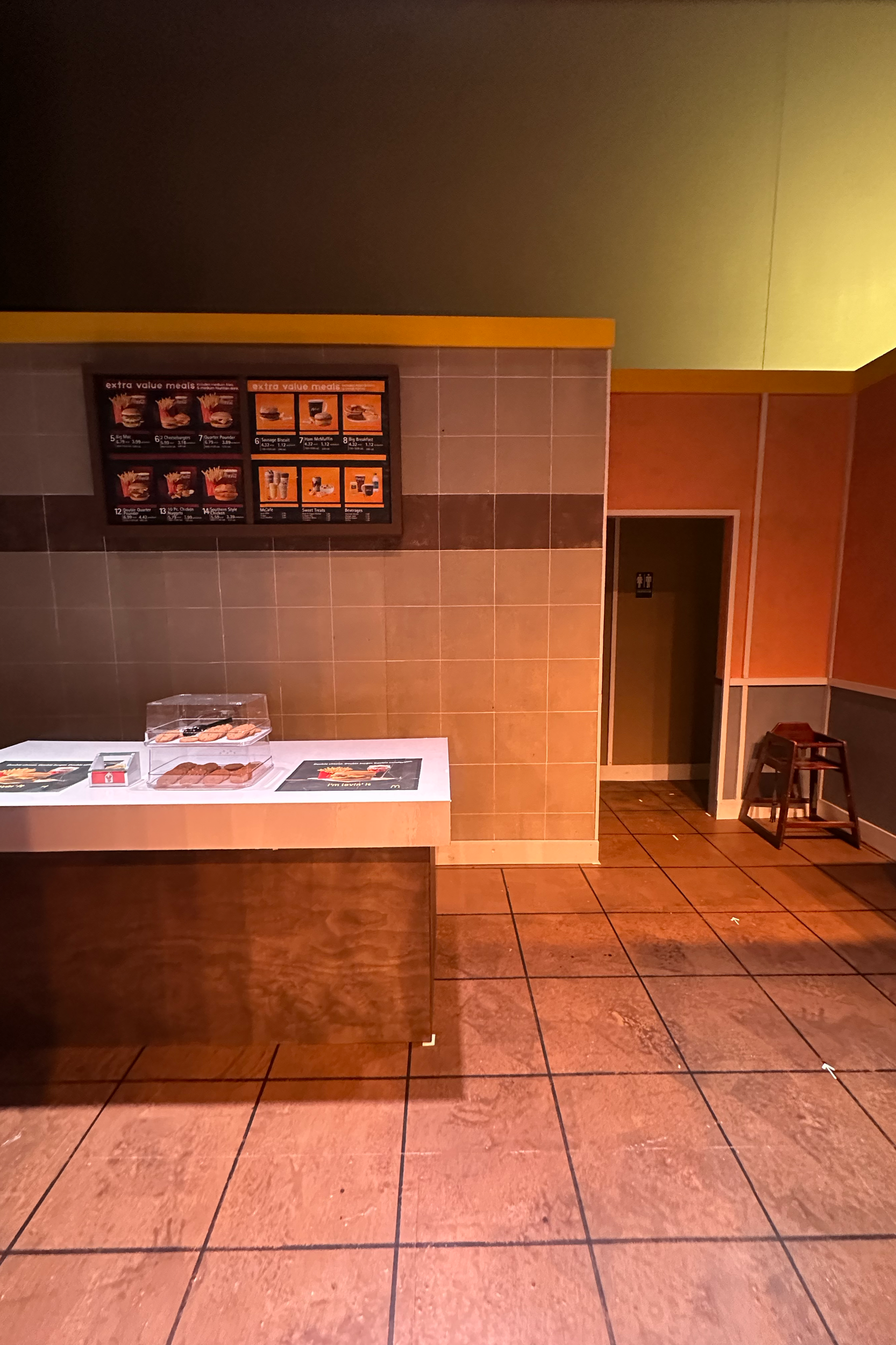 Empty interior of a fast-food restaurant with a white counter displaying pizza boxes, brown tiled wall, and menu board with pizza options in the background.