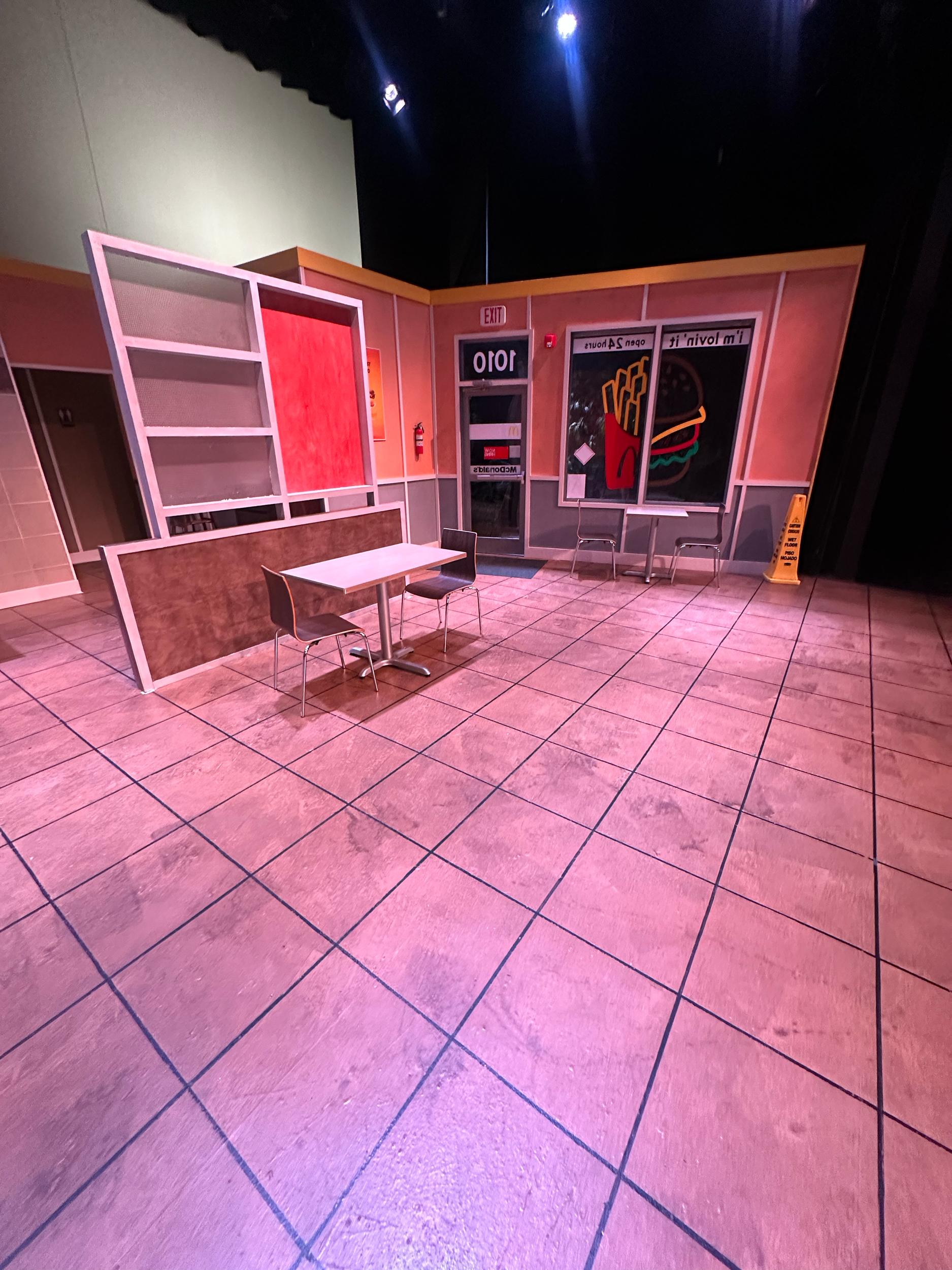 Empty table with three chairs in front of a colorful wall with neon signs and a door, illuminated by pink and purple lights.