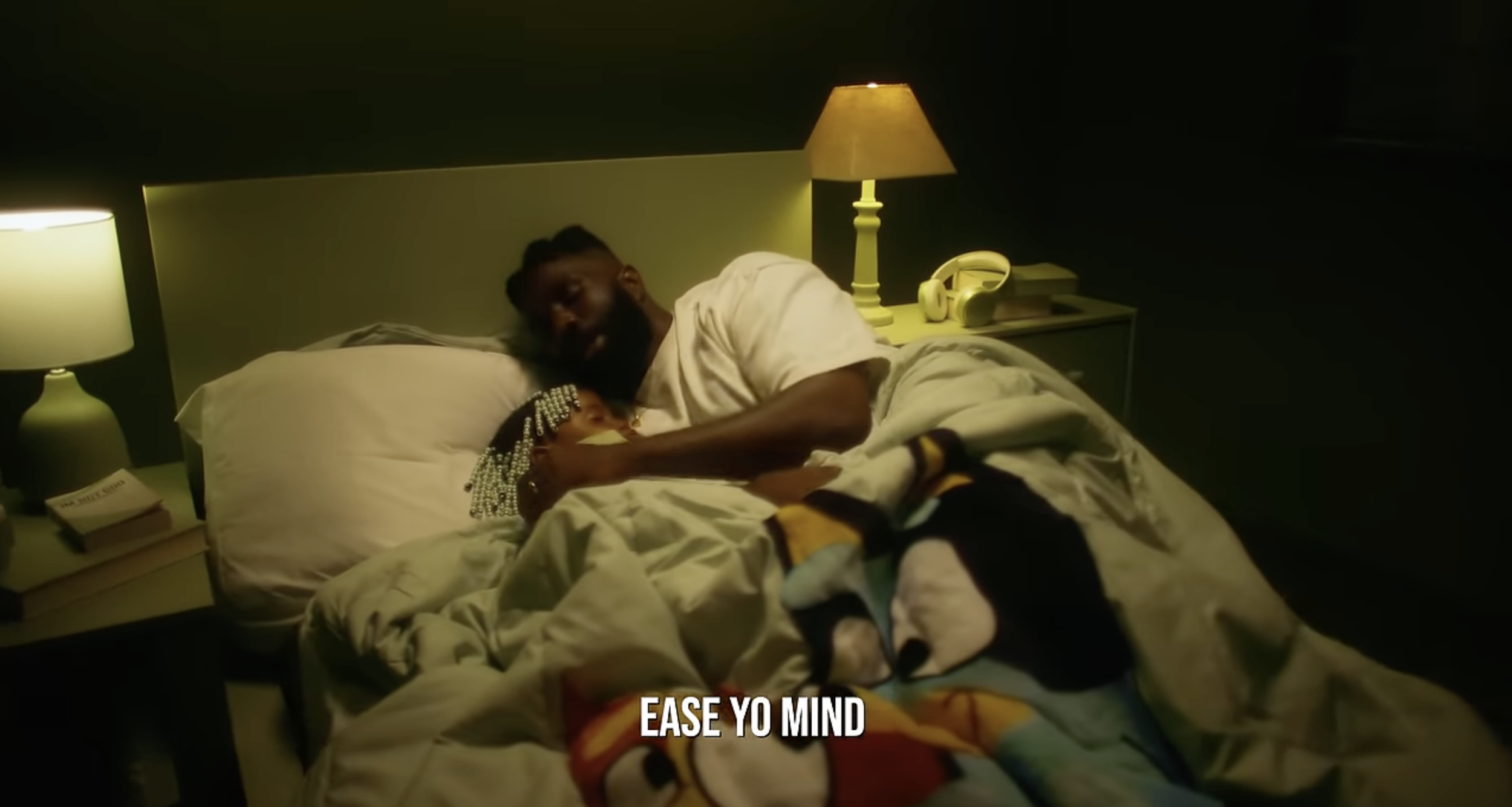 A man and a child lying in bed together, cuddling in a dimly lit bedroom with two yellow lamps on bedside tables.