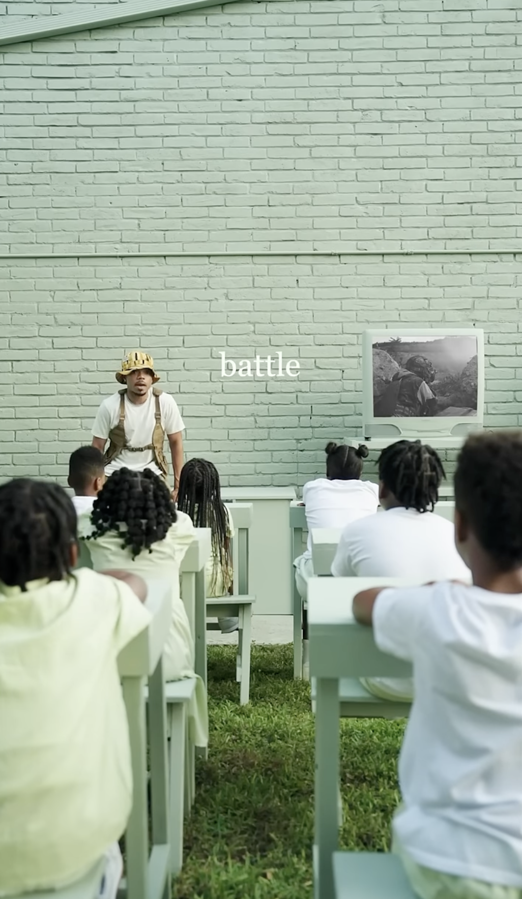A teacher in military gear standing in front of a classroom of young students outdoors, with a brick wall background and vintage television showing footage, captioned 'battle'.