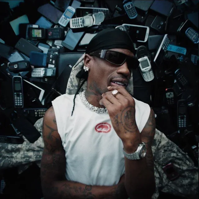 A man lying on a pile of older model cell phones, wearing sunglasses, a black head covering, a white sleeveless shirt, and jewelry including a diamond ring and watch.