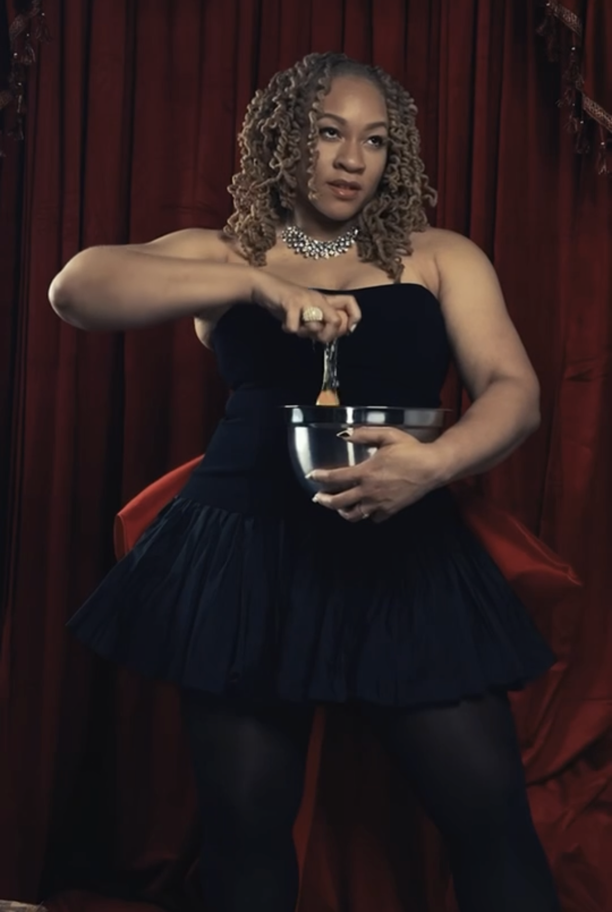 A woman with blonde curly hair in a strapless black dress, jewelry, and black tights, pouring liquid from a spoon into a silver bowl, sitting against a red curtain backdrop.