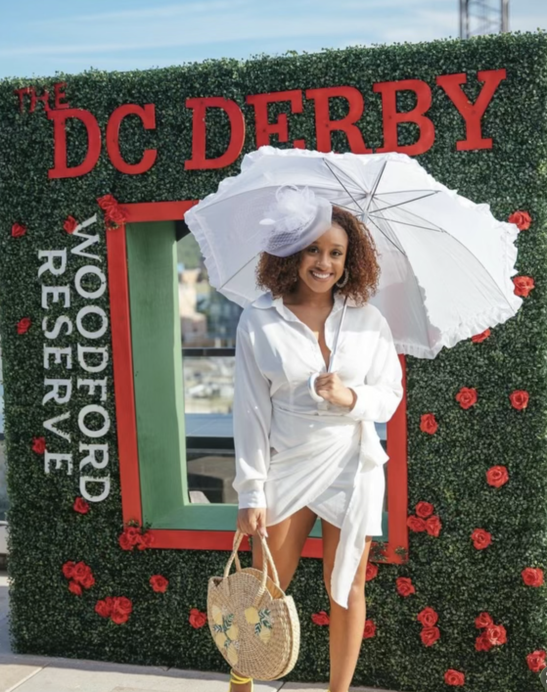 A woman in white dress holding a white umbrella and a straw bag, standing in front of a decorated hedge with red letters reading 'DC Derby' and 'The Pleasure of the Word Reserve' at the event.