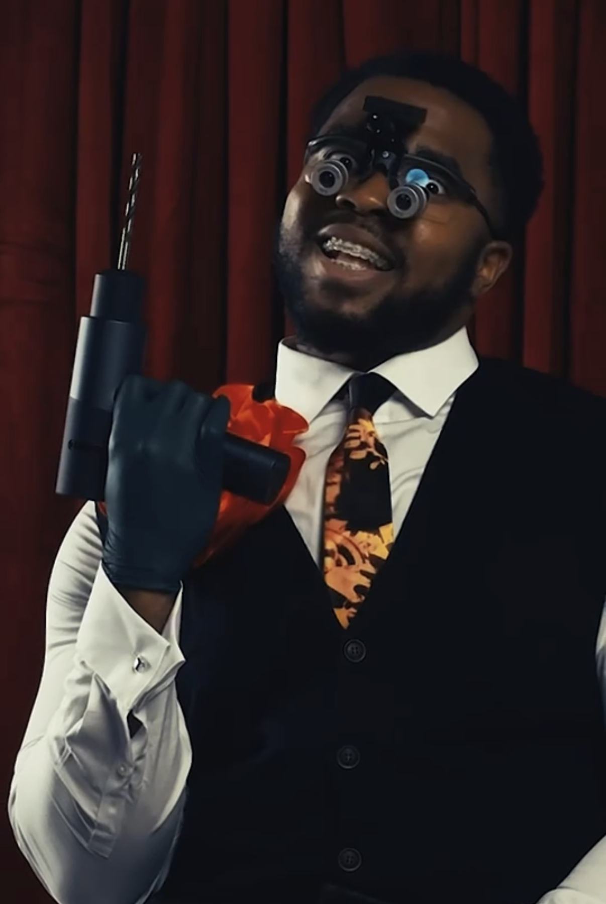 A man wearing black gloves and a suit with a colorful tie, holding a gun, with a headband that has small cameras or lenses, standing in front of a wooden background.